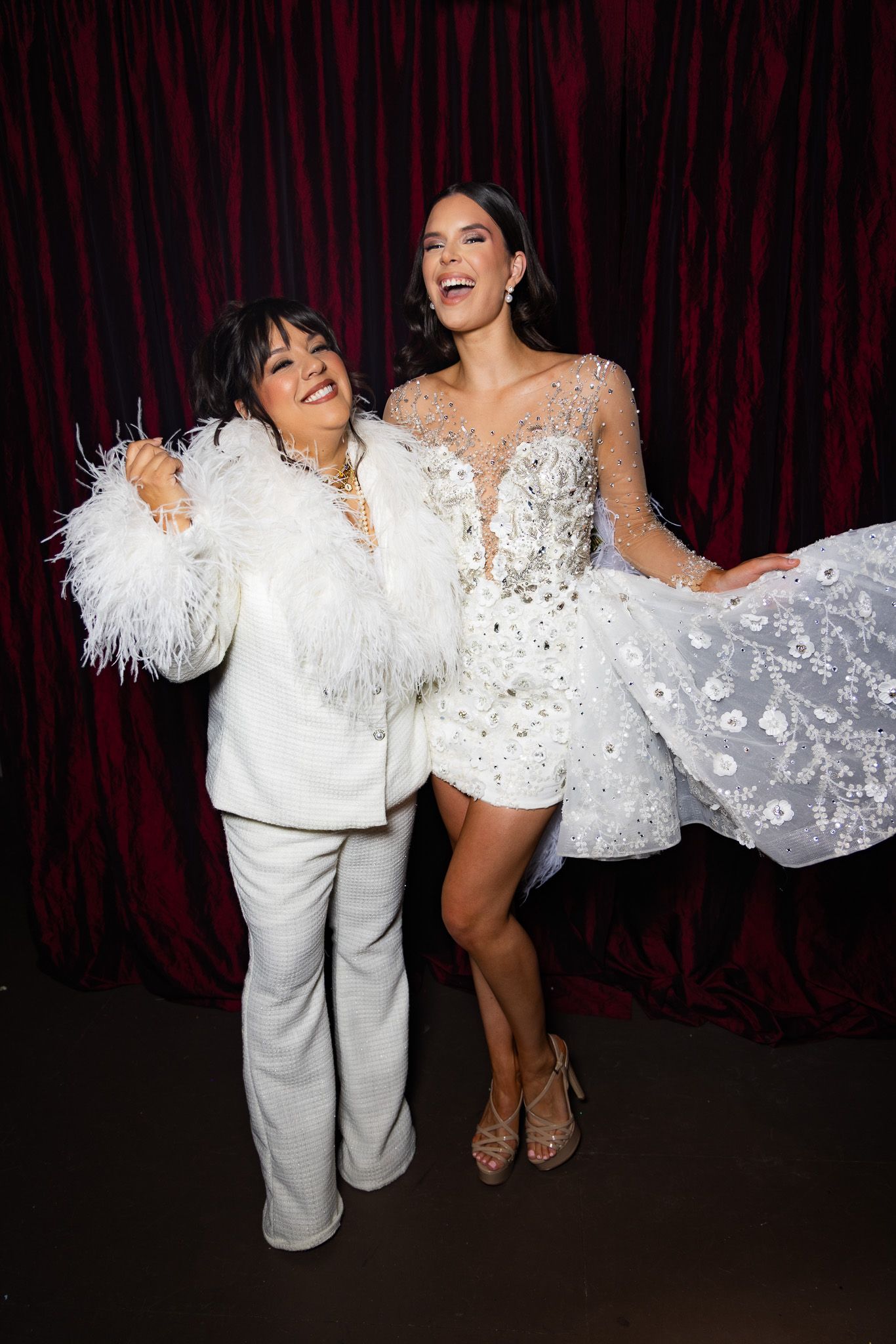 Two figures in glamorous white outfits pose and laugh together against a dark red curtain backdrop.