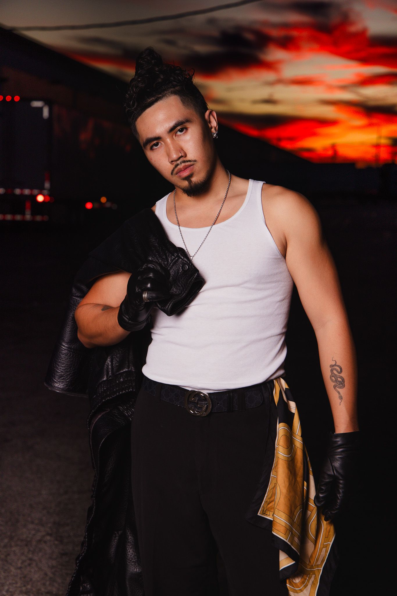 Dramatic portrait against a fiery sunset sky wearing a white tank top and black leather gloves.