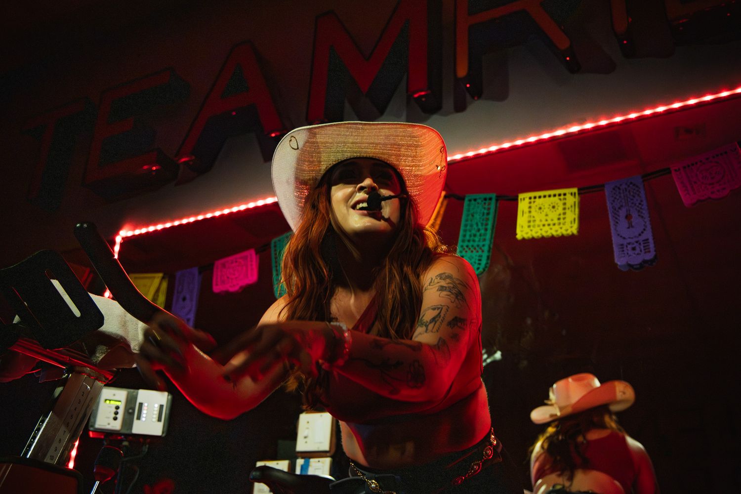 Tattooed female drummer in white cowboy hat performing energetically under neon lights with papel picado.