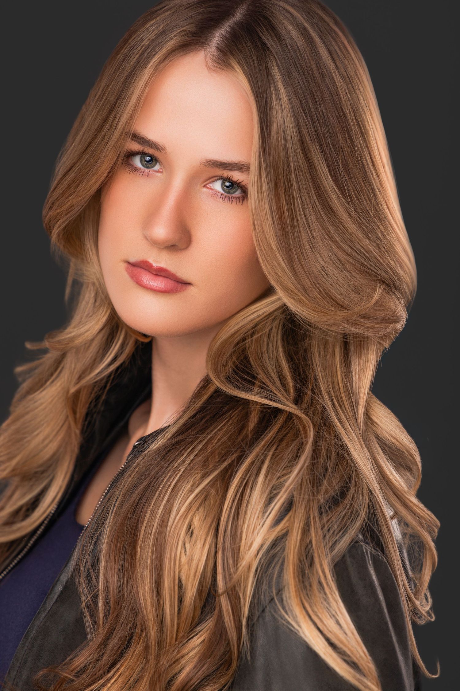 Professional beauty portrait with wavy caramel blonde hair against dark background.