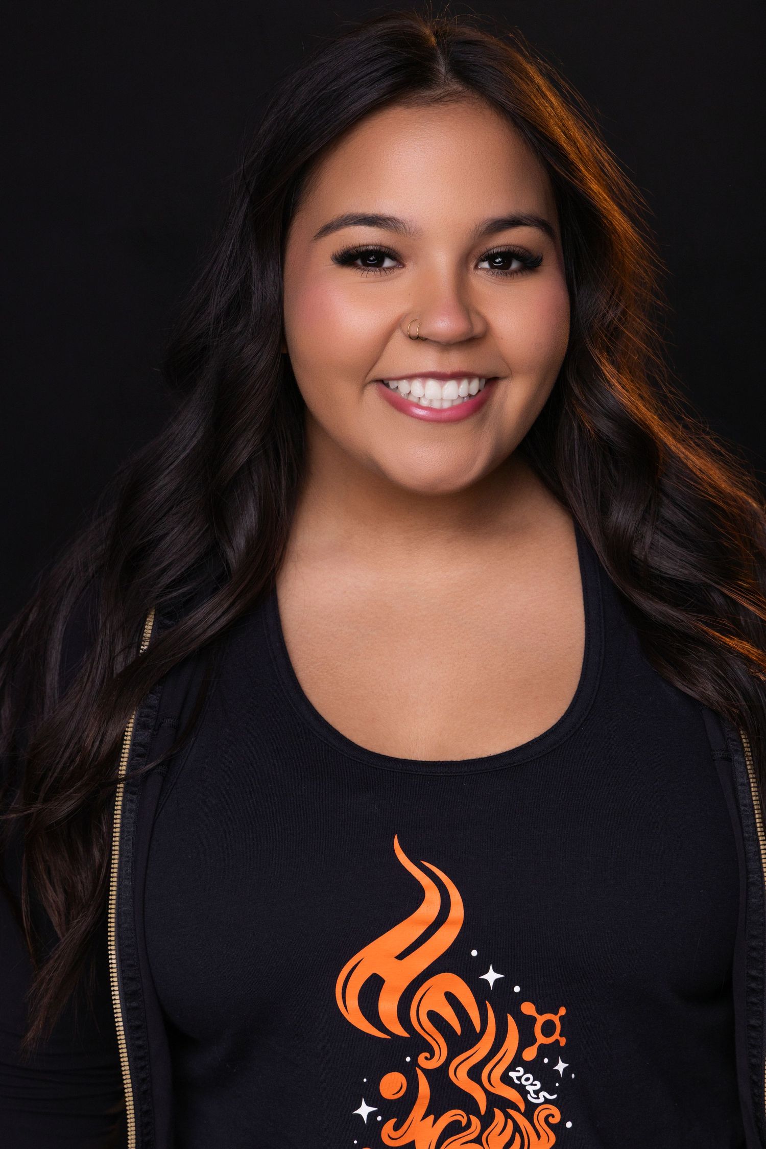 Studio portrait of a smiling person with long dark hair wearing a black top with orange decorative design against dark background.