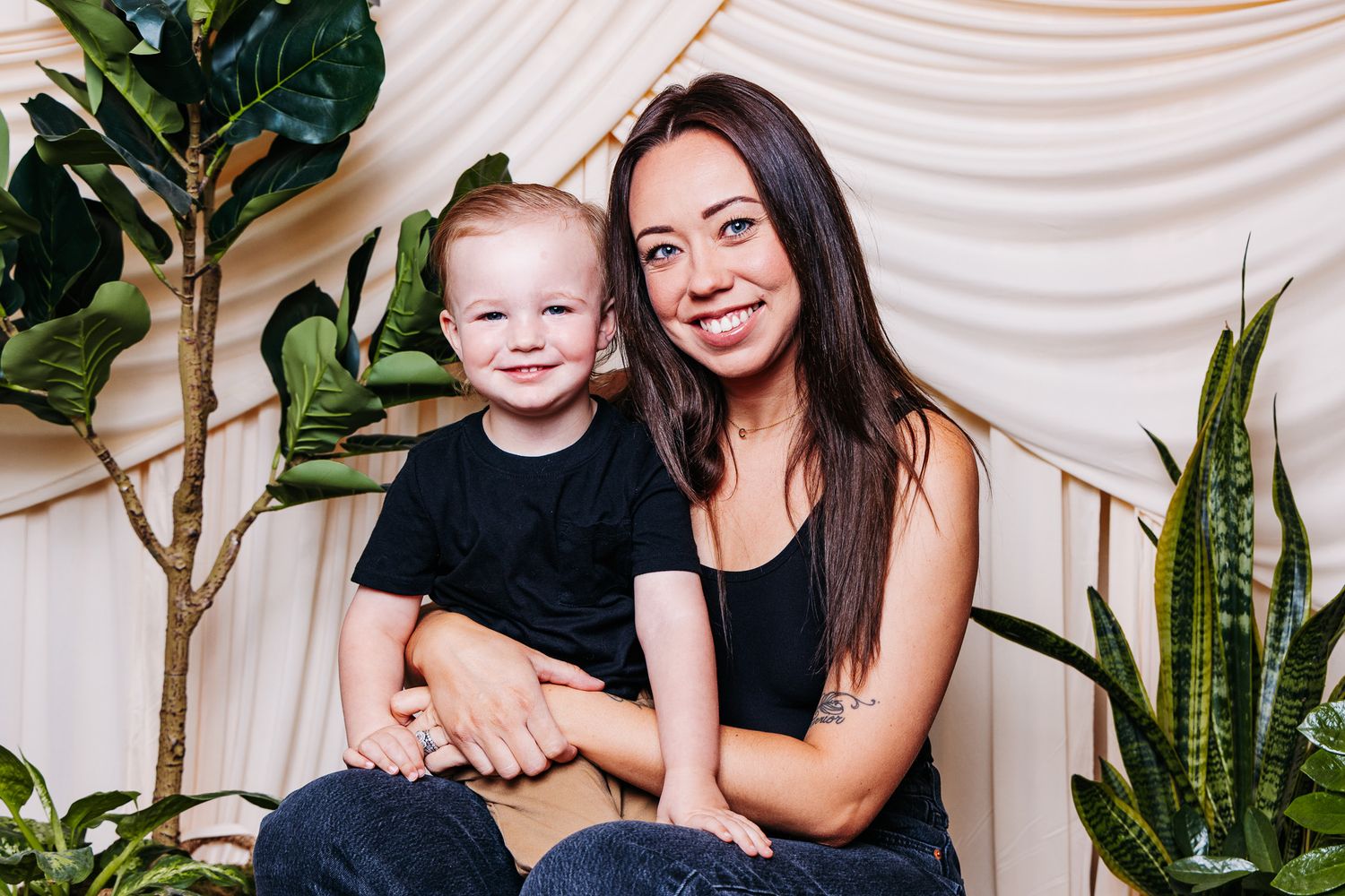 A smiling woman and young red-haired boy in black outfits pose together among green plants on a white backdrop.