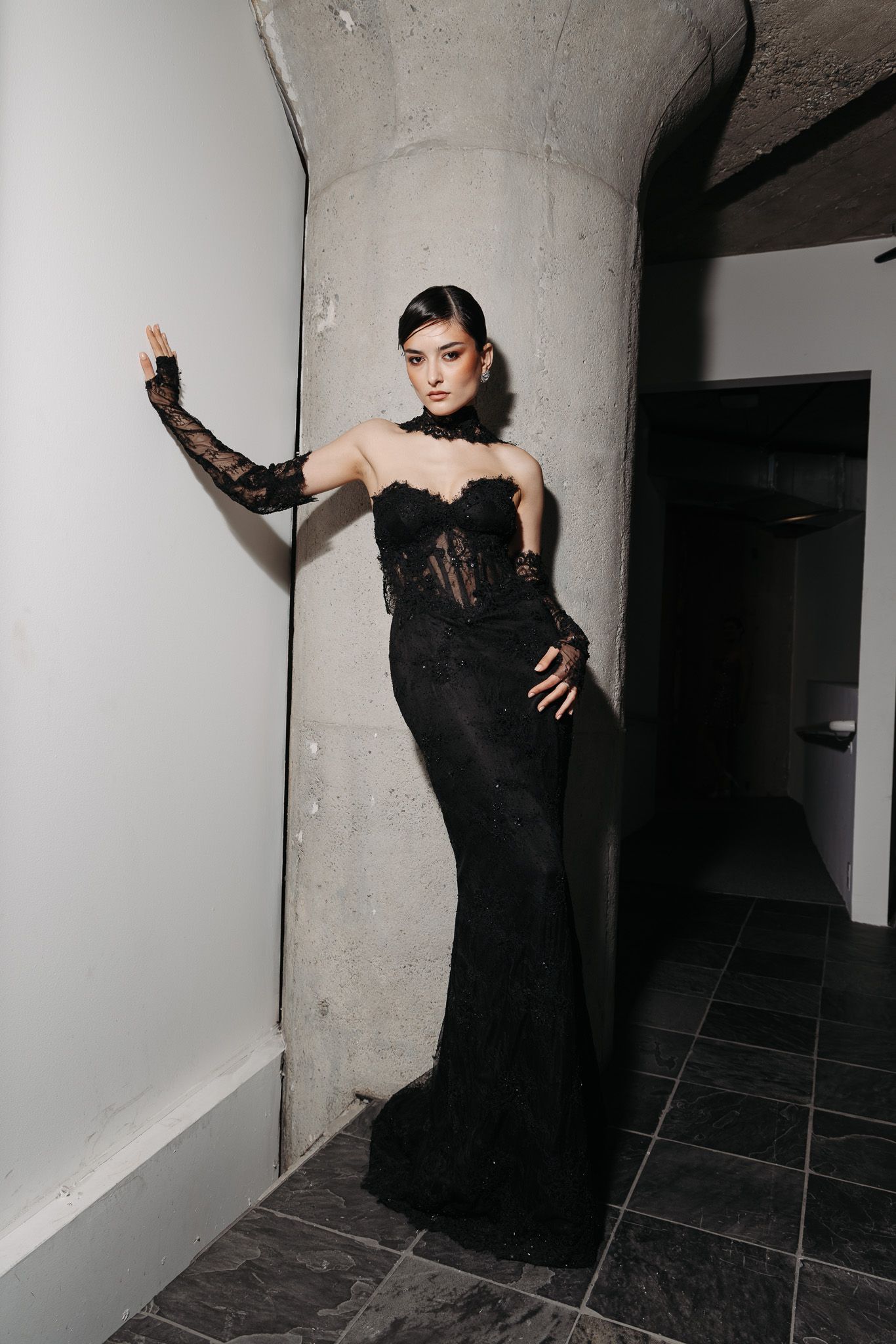 Model poses in elegant black lace evening gown with sheer details against industrial concrete wall.