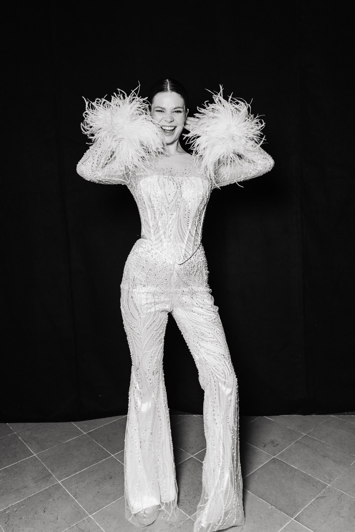 Performer in sparkling white jumpsuit with dramatic feather details strikes playful pose against black backdrop.