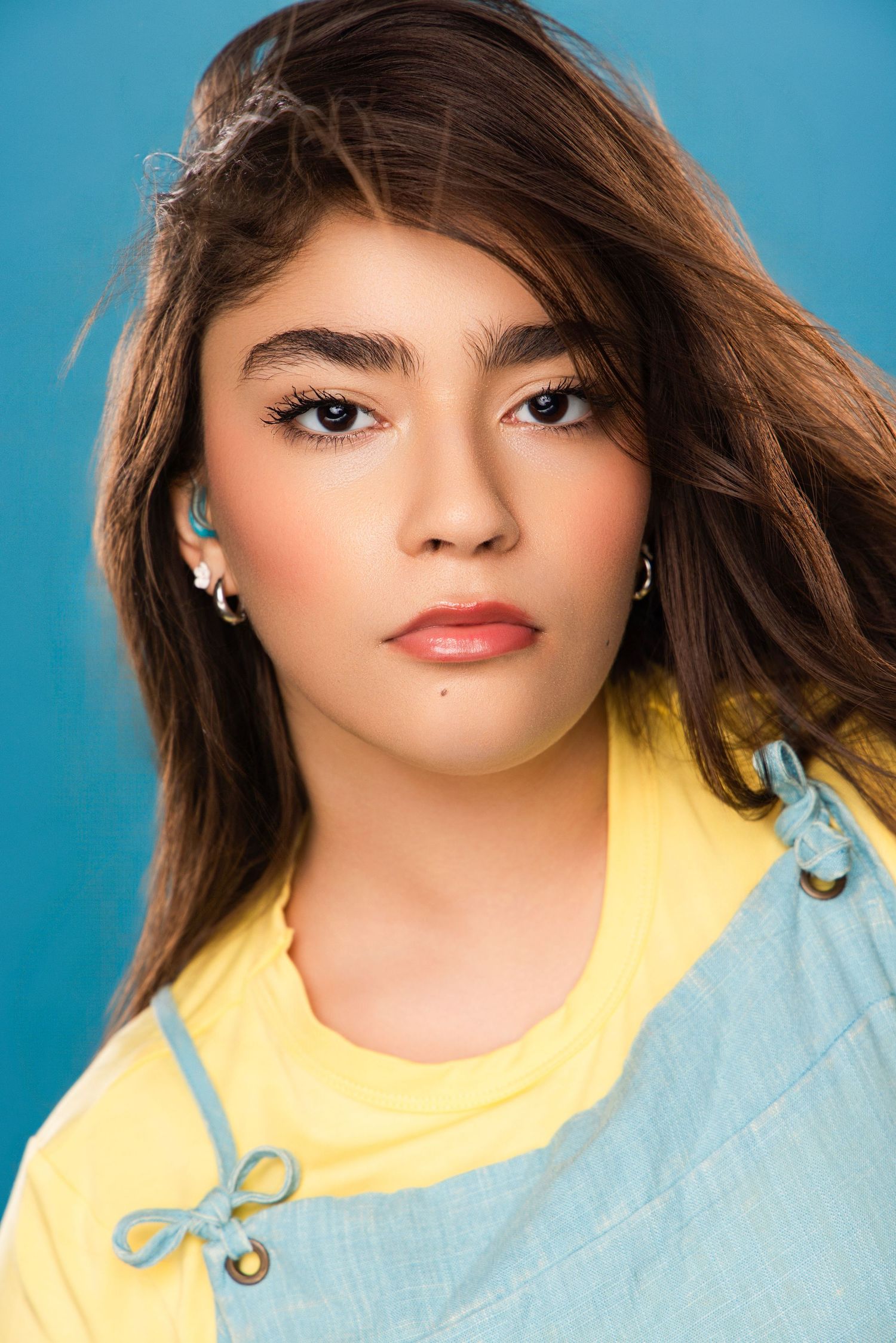 Fashion portrait against blue background wearing yellow and light blue casual top with flowing brown hair.