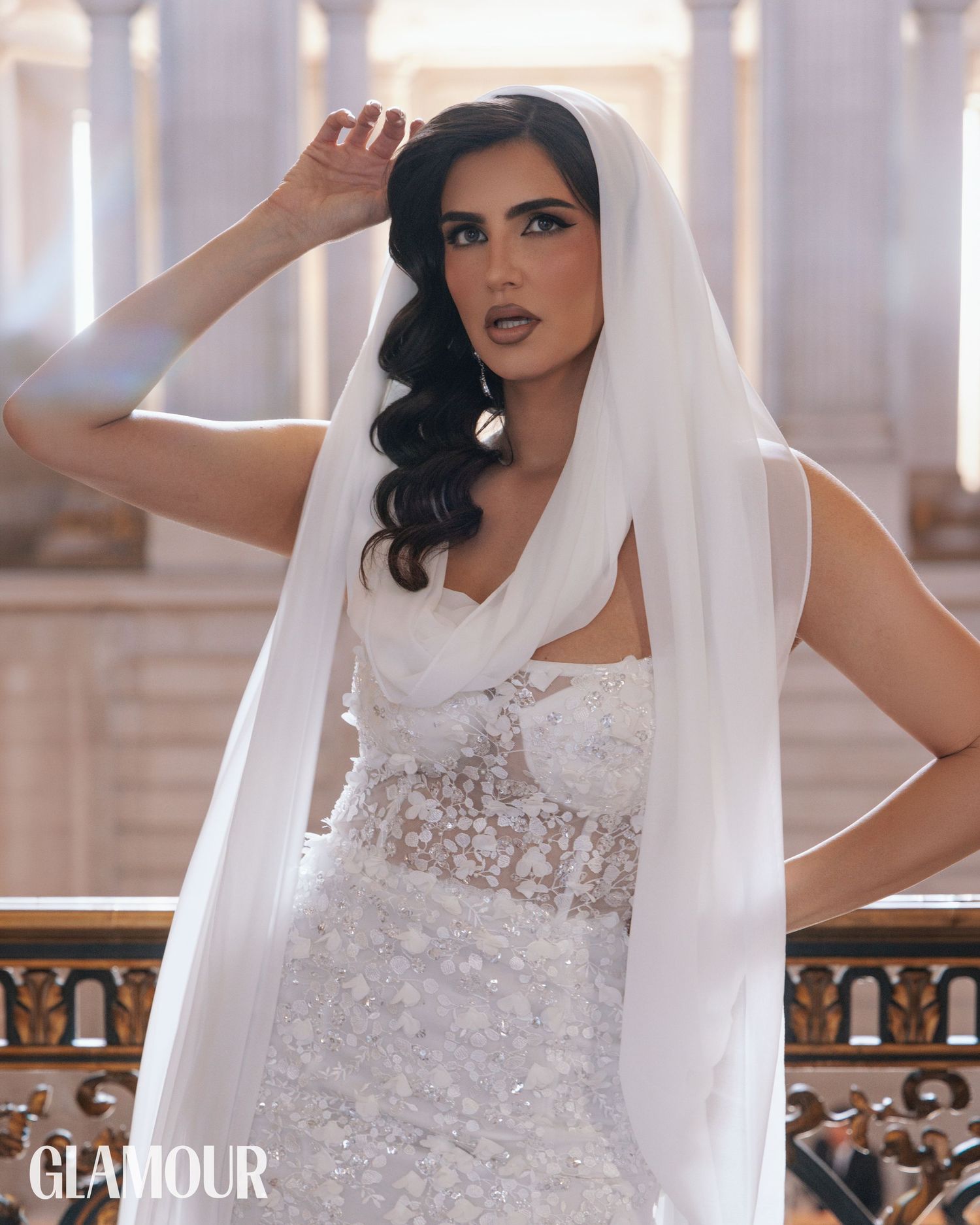 A bride in a detailed lace wedding gown and flowing veil poses elegantly against a backdrop of classical architecture.