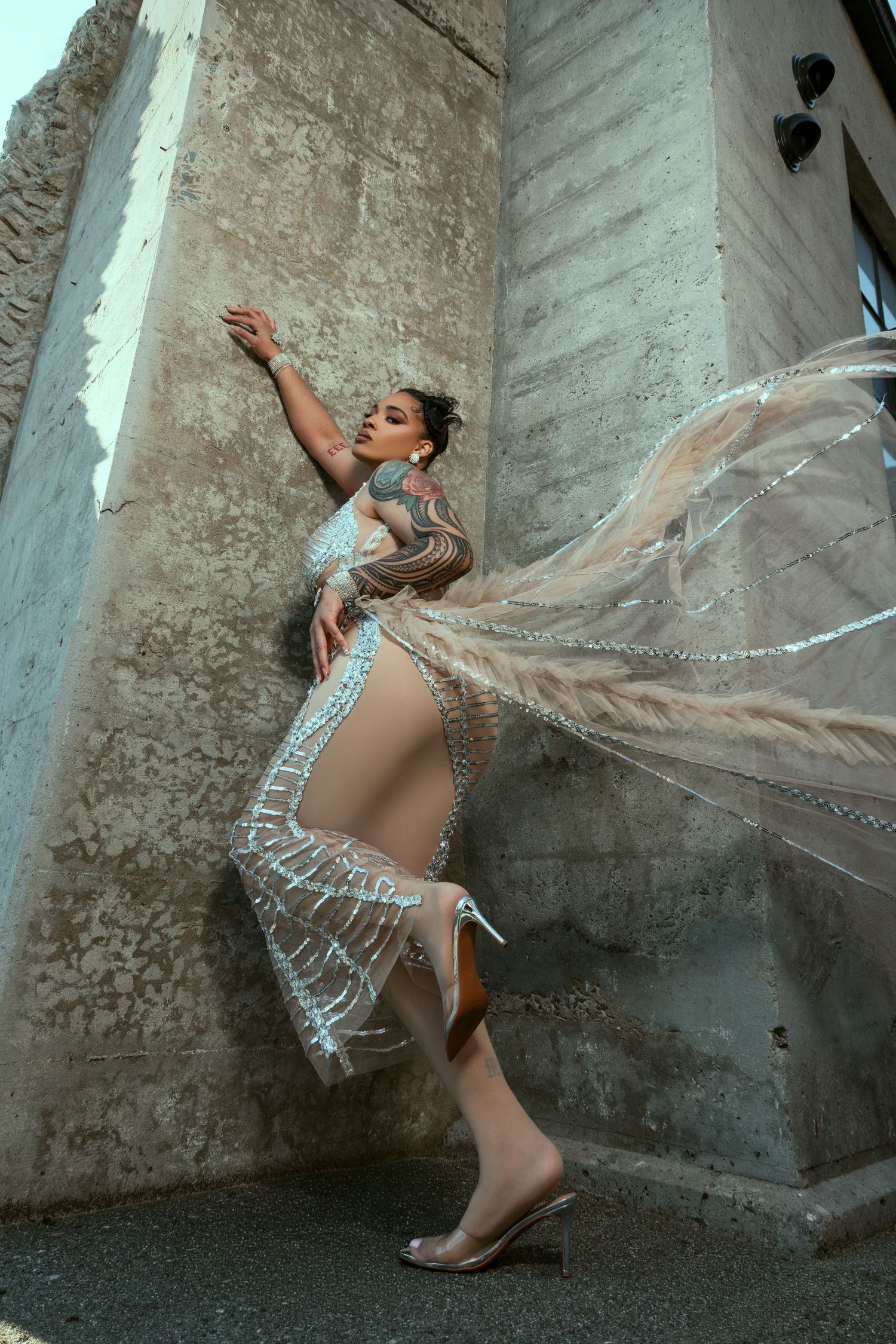 Graceful dancer in a beige bodysuit and flowing white cape performs dynamic movements against a textured stone wall.