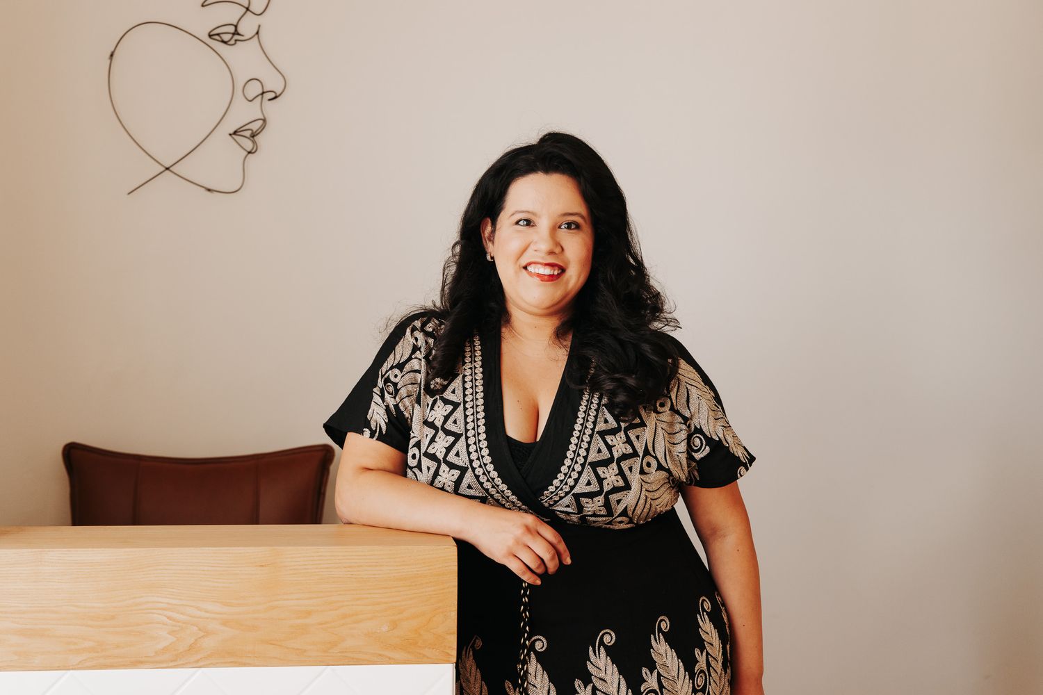 A person wearing a black and white floral dress stands against a light colored wall in a home setting.