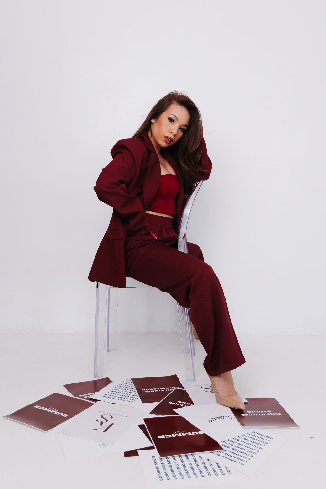 Woman in burgundy suit sitting on clear chair surrounded by scattered papers on white background.