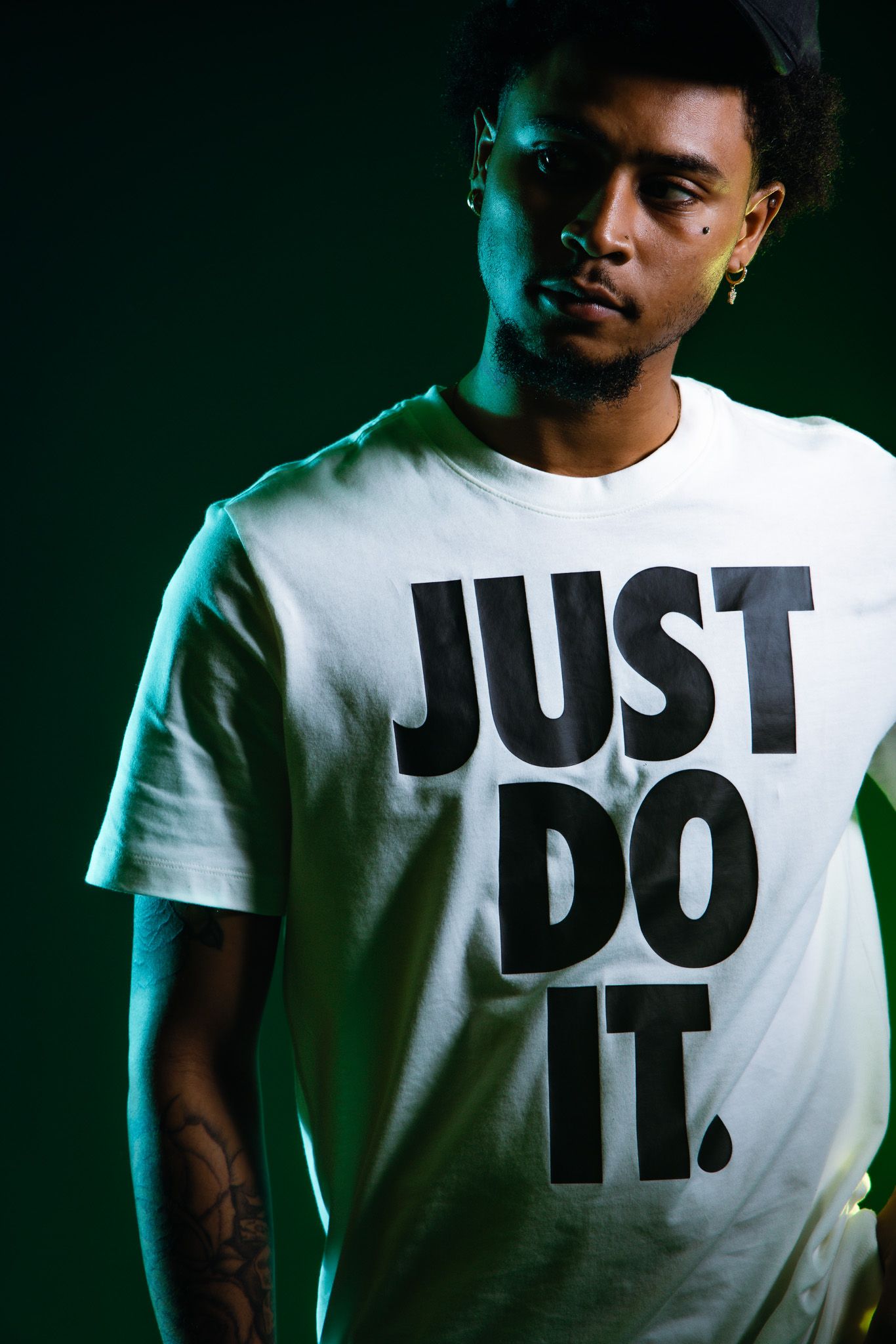 Young man wearing white Nike Just Do It t-shirt with arm tattoo under green studio lighting.