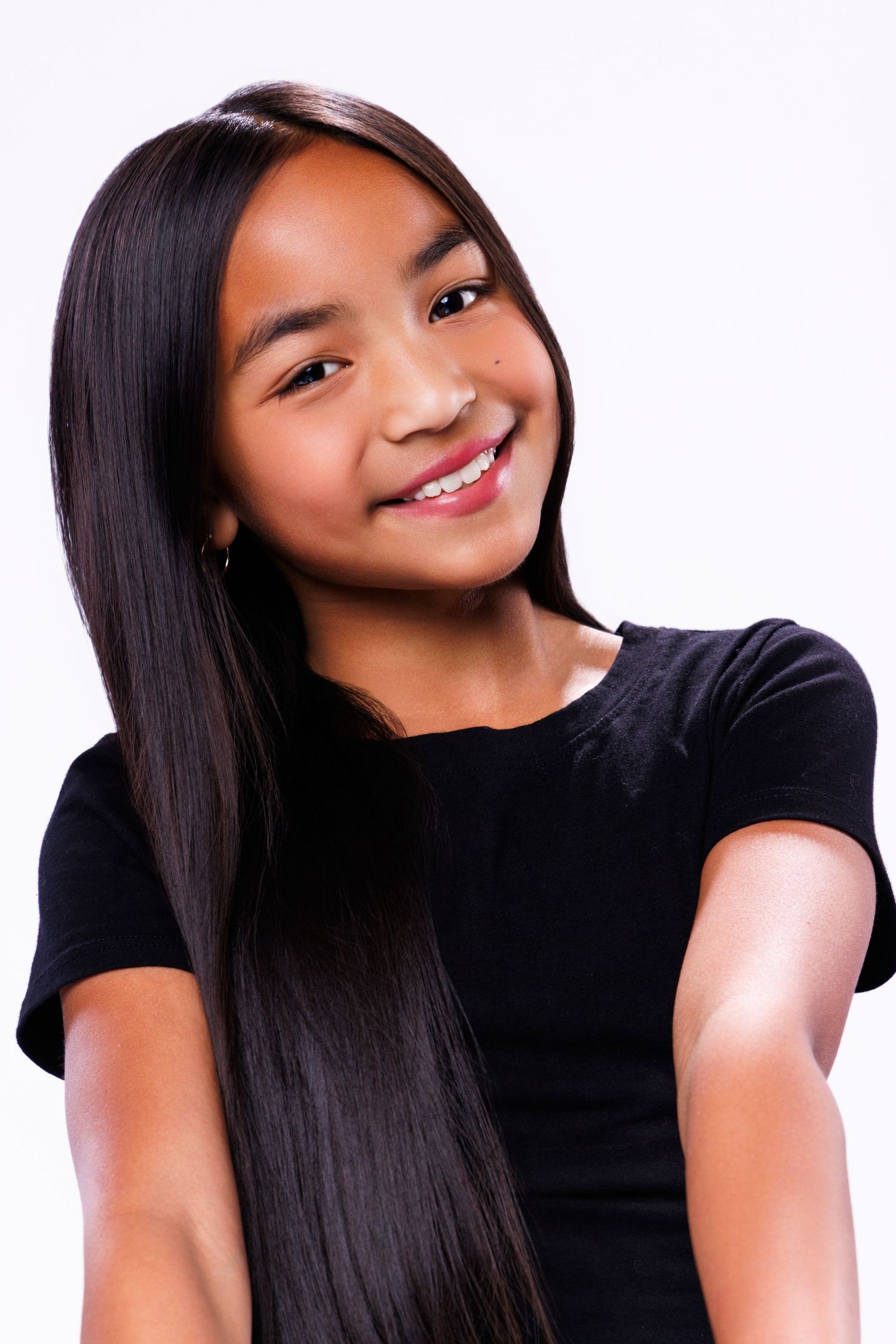Young girl with long straight black hair smiling, wearing a black top against a white background.