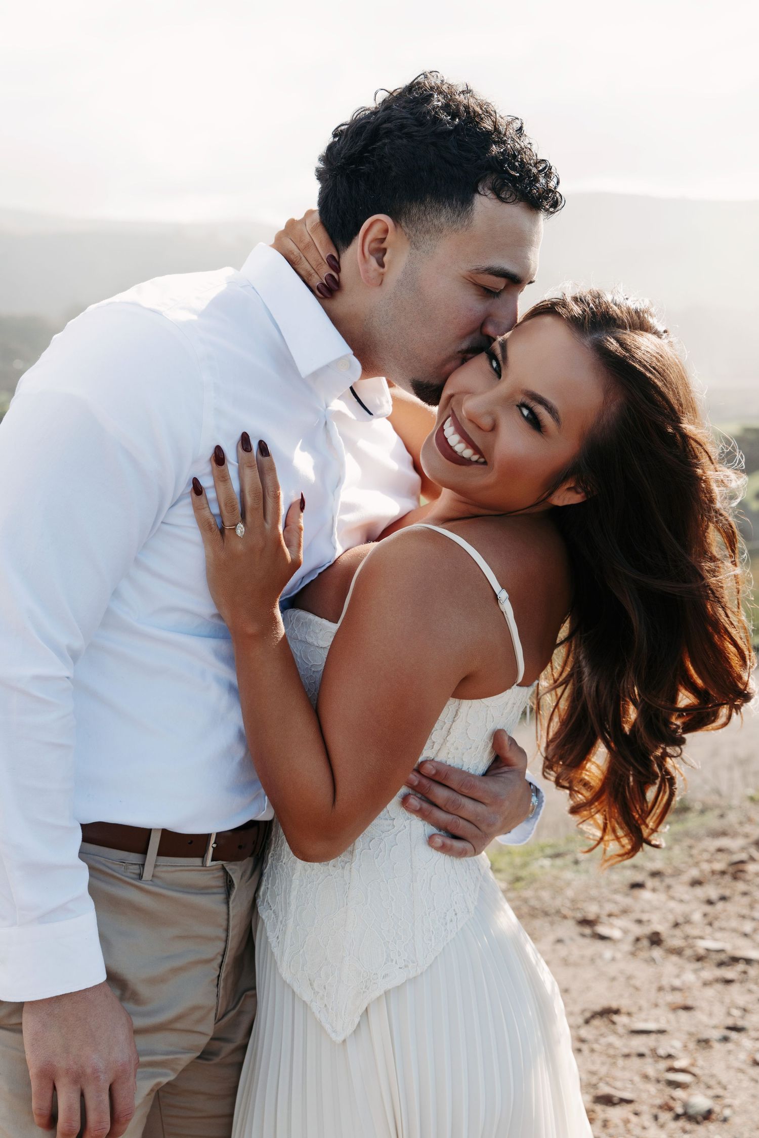 A couple in white and beige attire share intimate moments on a beach during a romantic photoshoot.