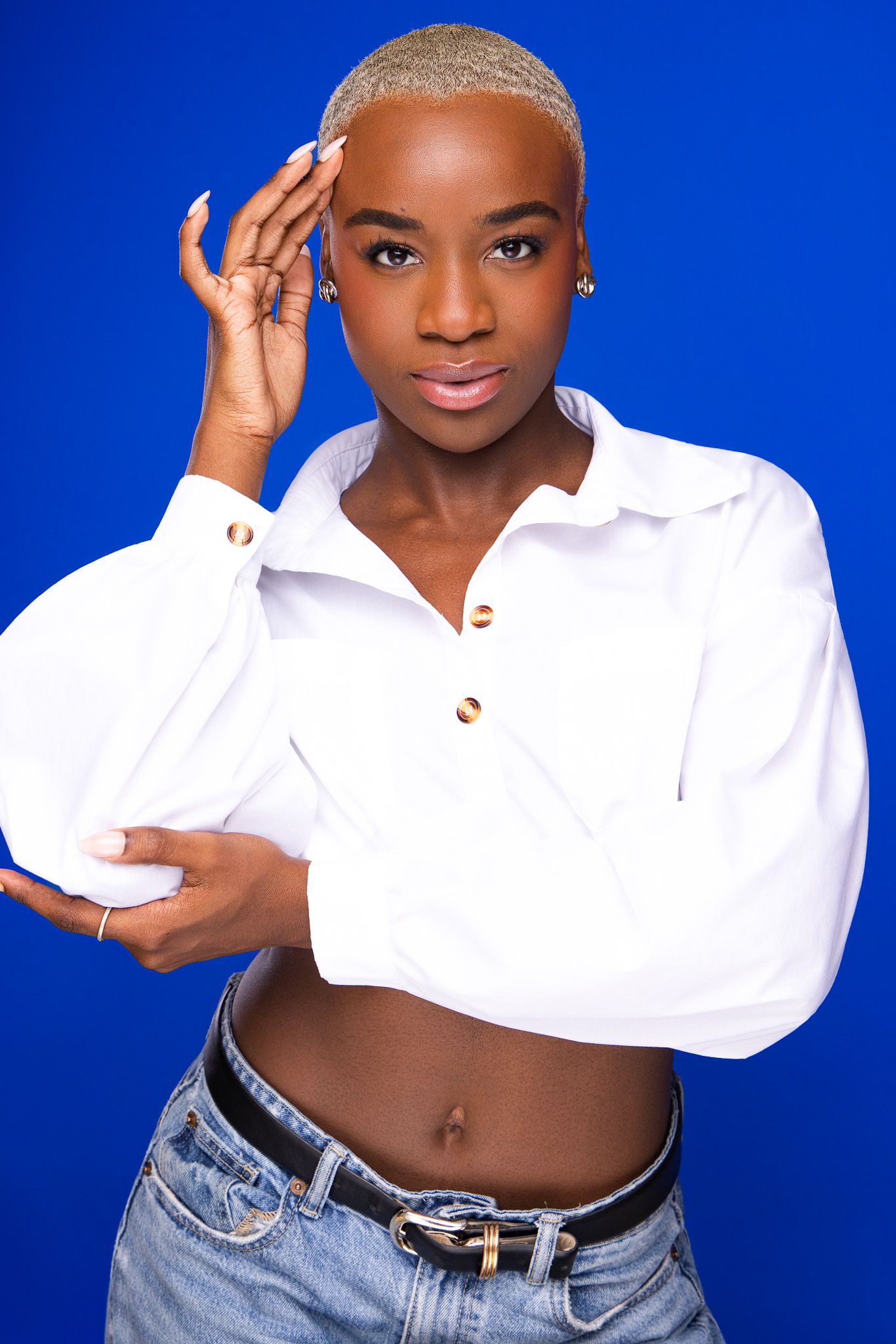 Young Black woman with blonde buzz cut wearing white crop top and jeans poses confidently against blue background.