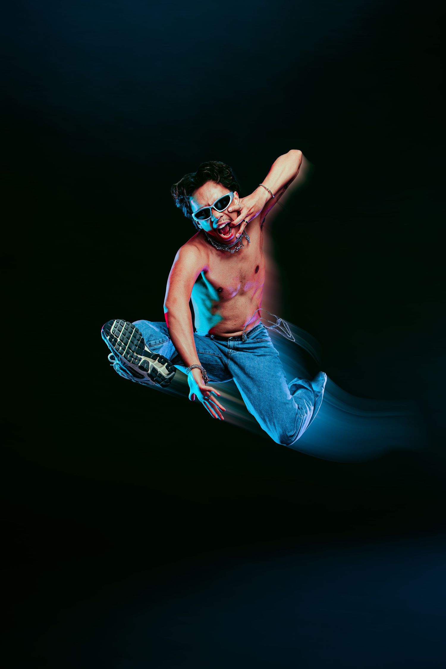 Shirtless male dancer jumping mid-air in blue jeans and sunglasses under neon blue light on dark background.