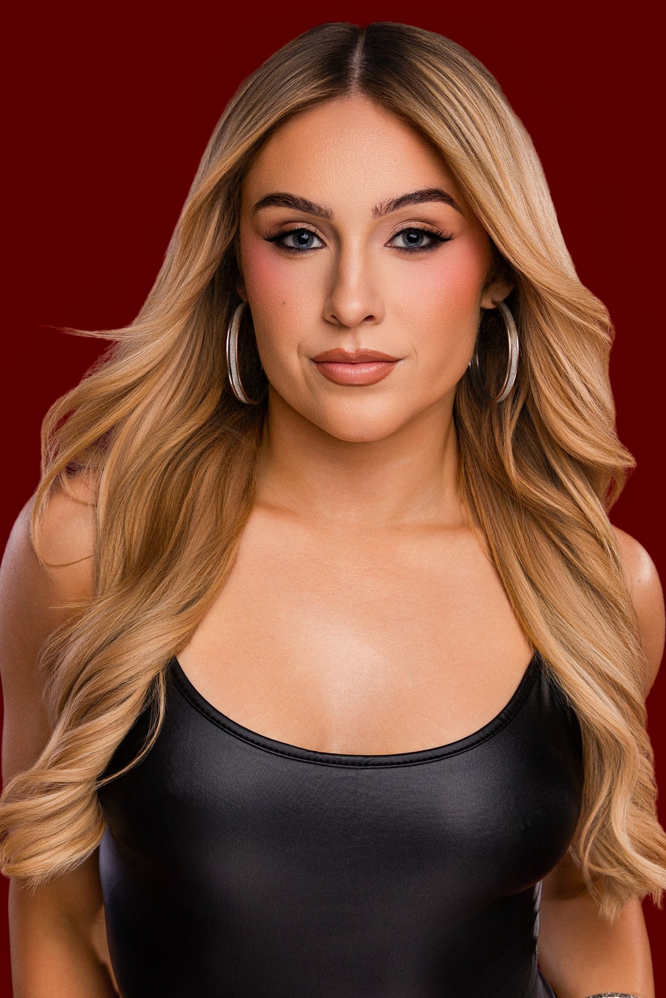 Young woman with long blonde wavy hair wearing a black leather top and hoop earrings against a red background.