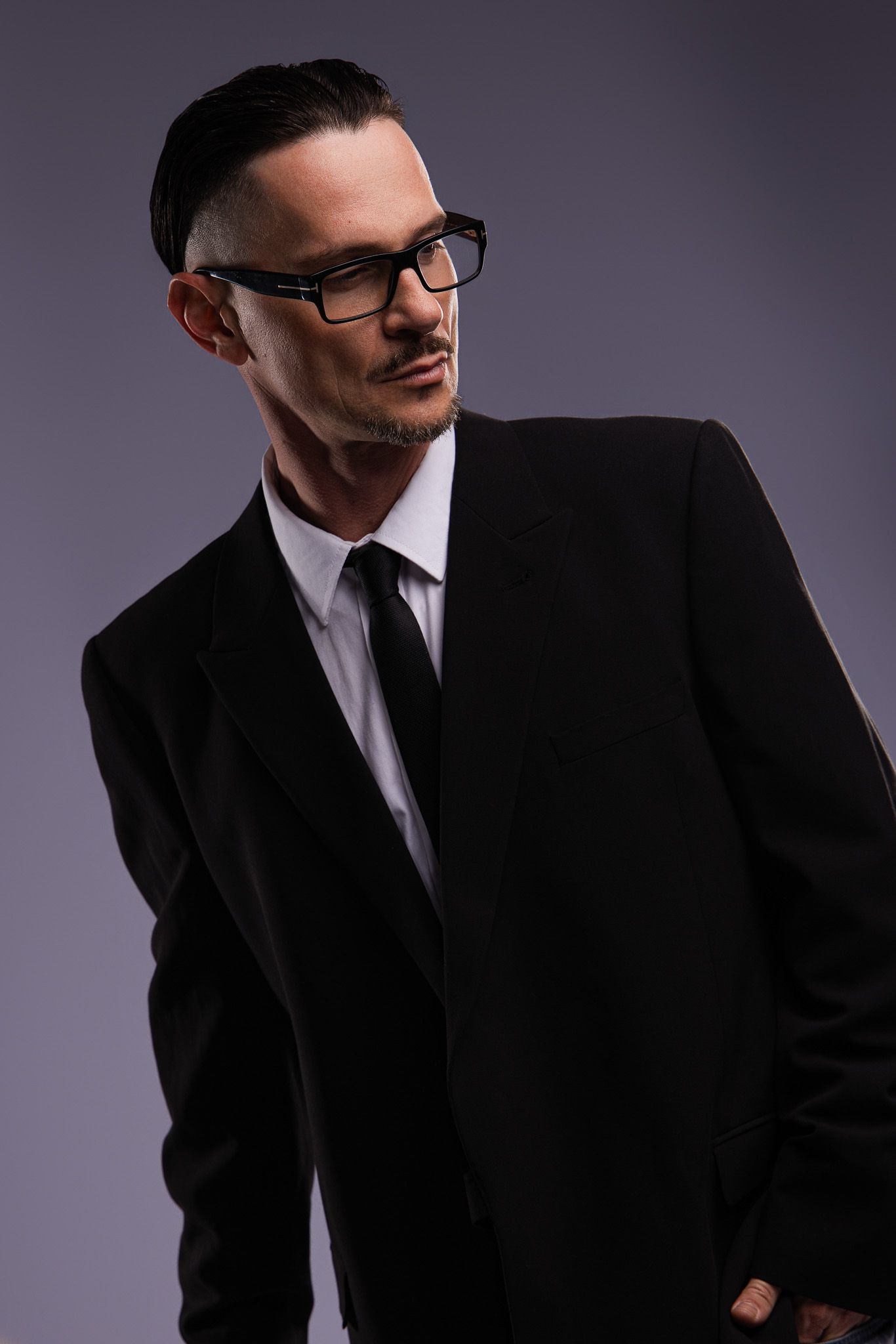 Man in black suit, white shirt, and tie wearing glasses, posing confidently against a gray background.