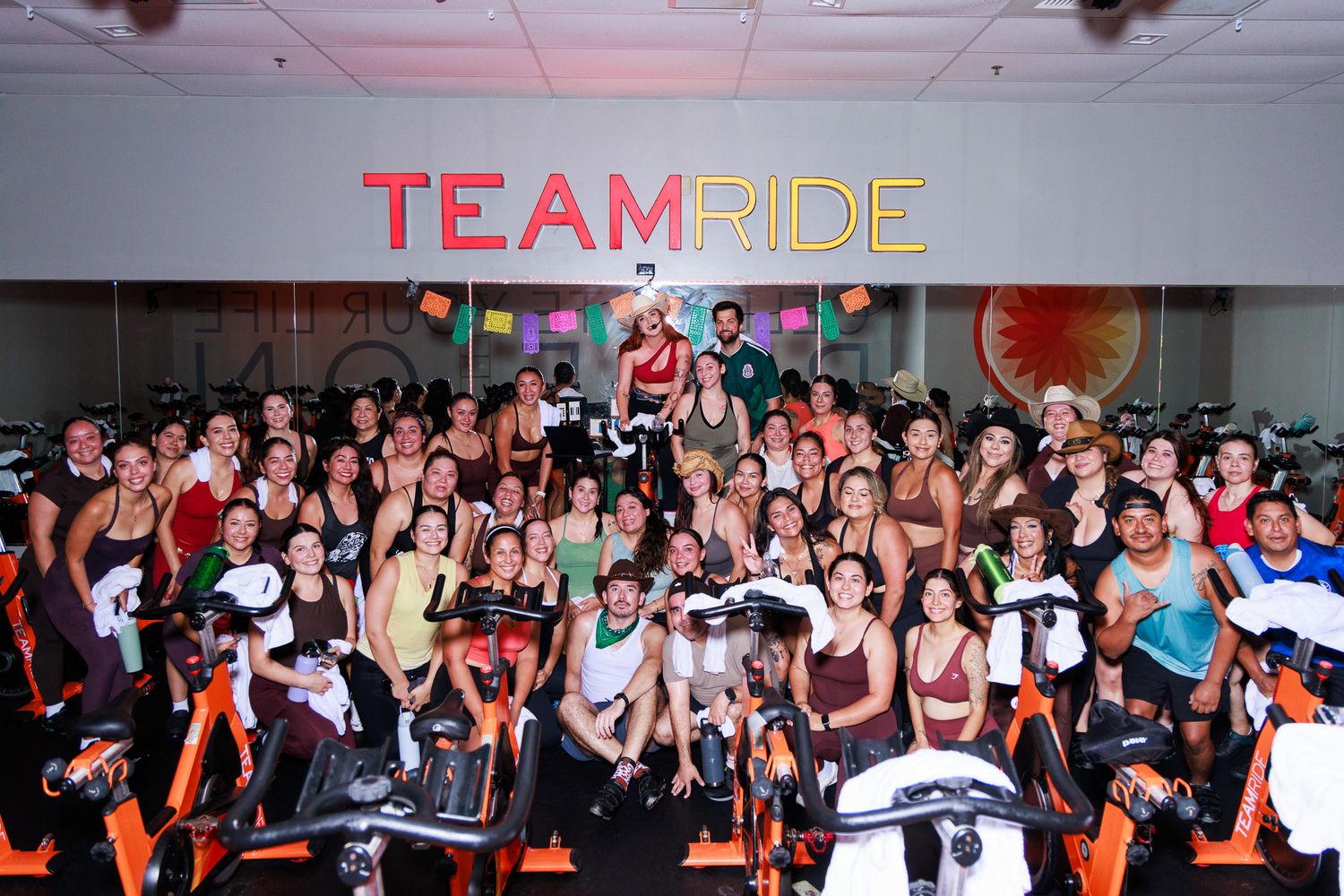 Large group of cyclists posing together in a Team Ride indoor cycling studio with orange and white decor.