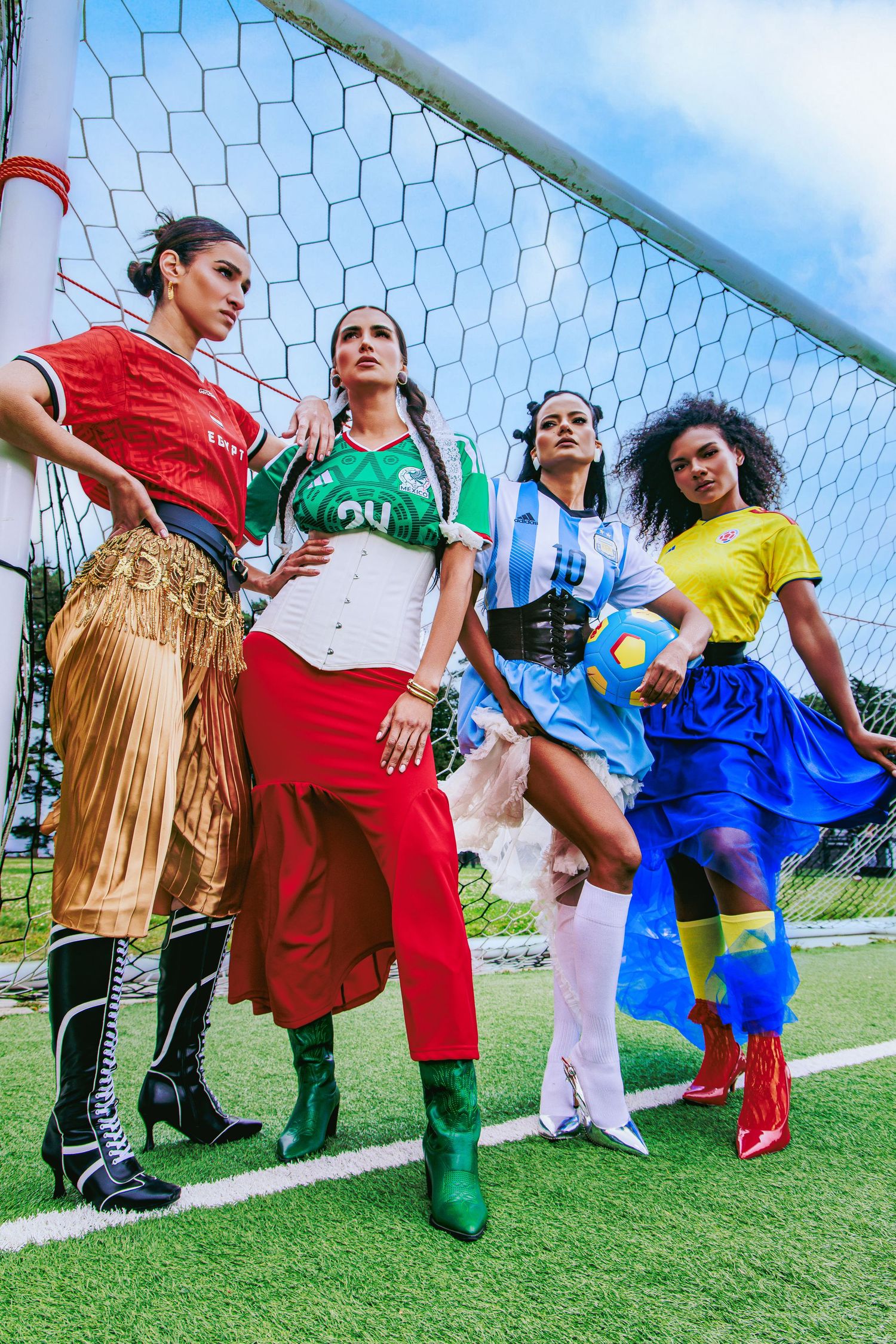 Four diverse women in vibrant soccer-inspired fashion pose confidently at a football goal on a green field.
