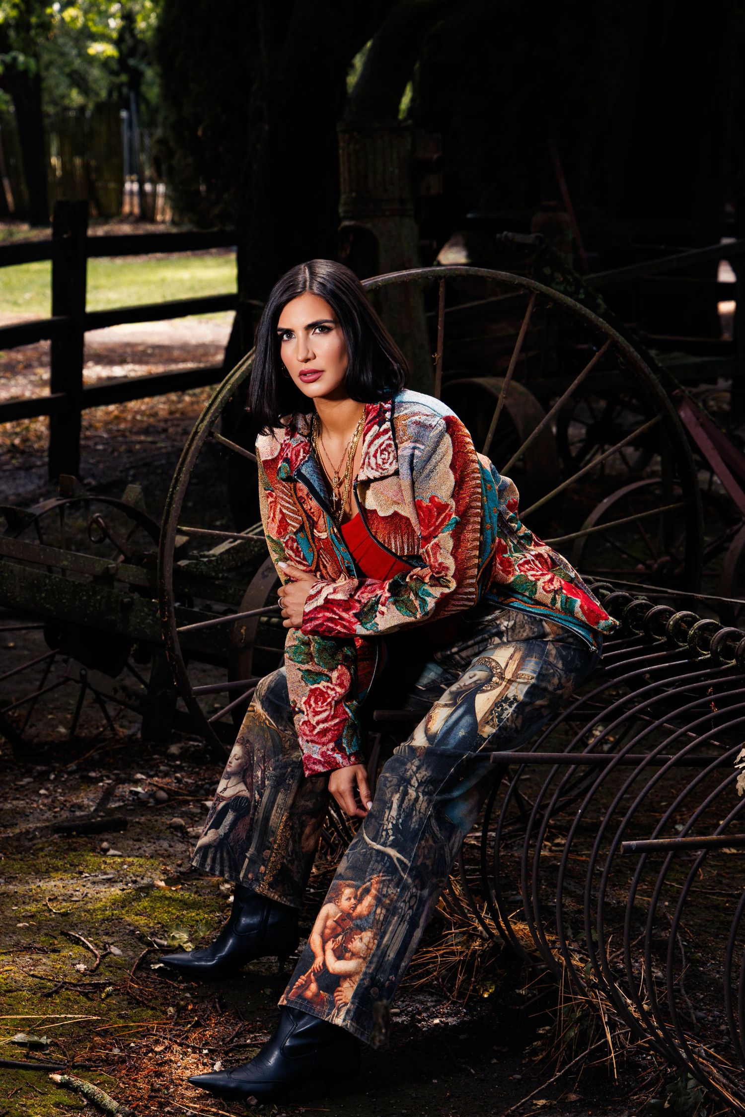 Woman in floral pantsuit and black boots posed on vintage farm equipment with rustic wooden fence backdrop.