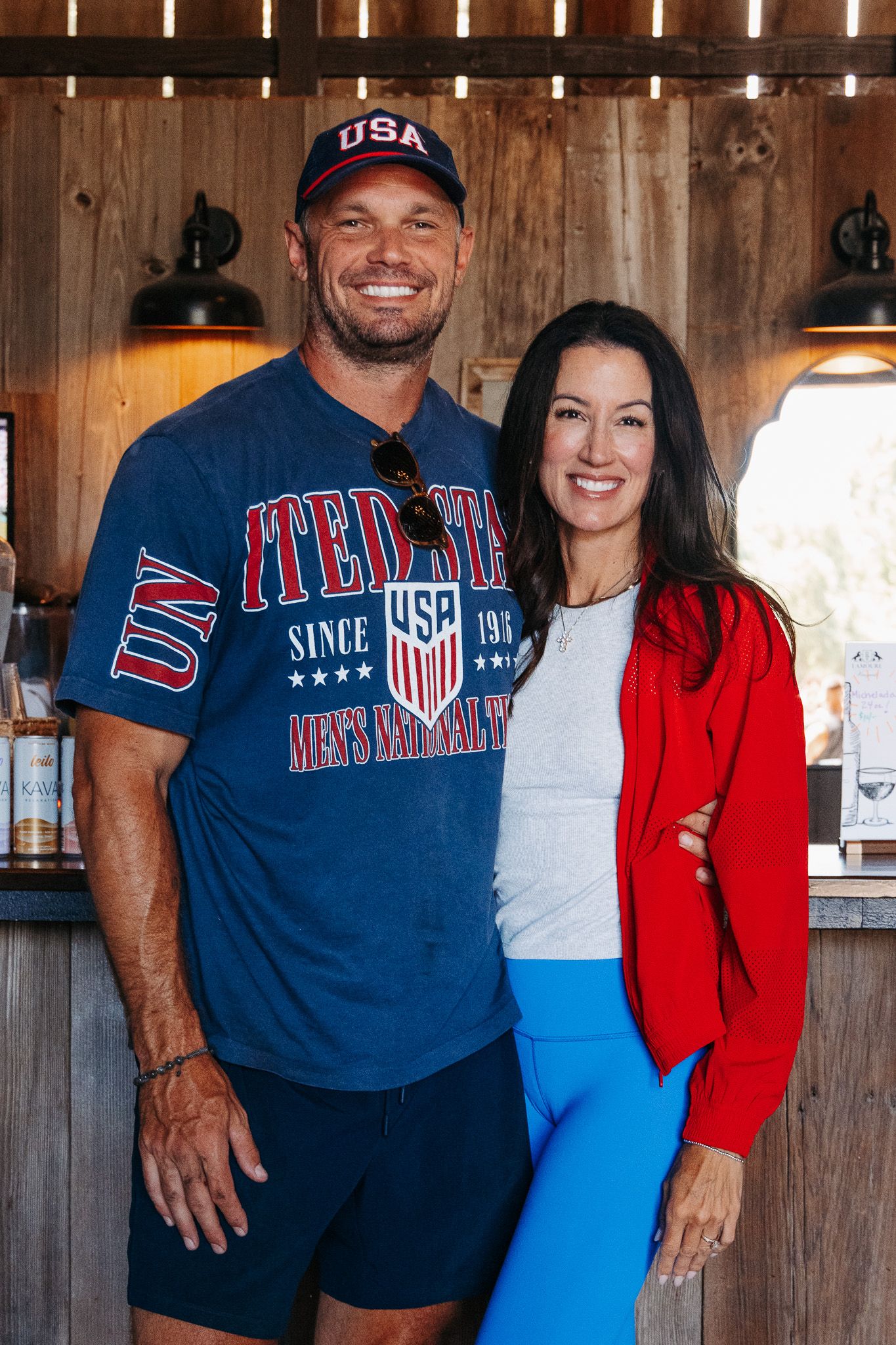 Smiling couple posing indoors, man in USA navy blue t-shirt and cap, woman in red jacket and blue leggings.