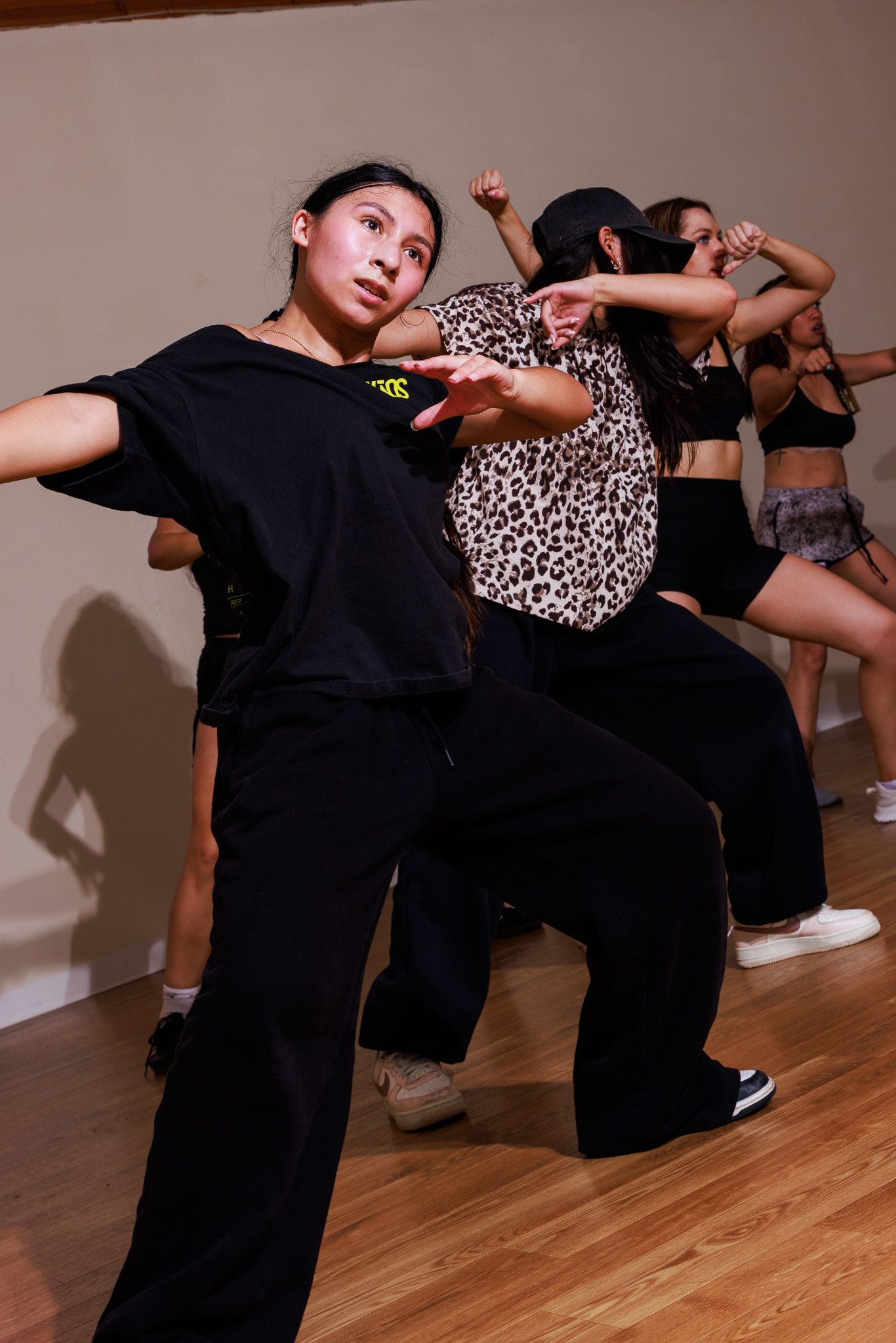 Young dancers practice hip-hop choreography in a dance studio, wearing casual black outfits on a hardwood floor.
