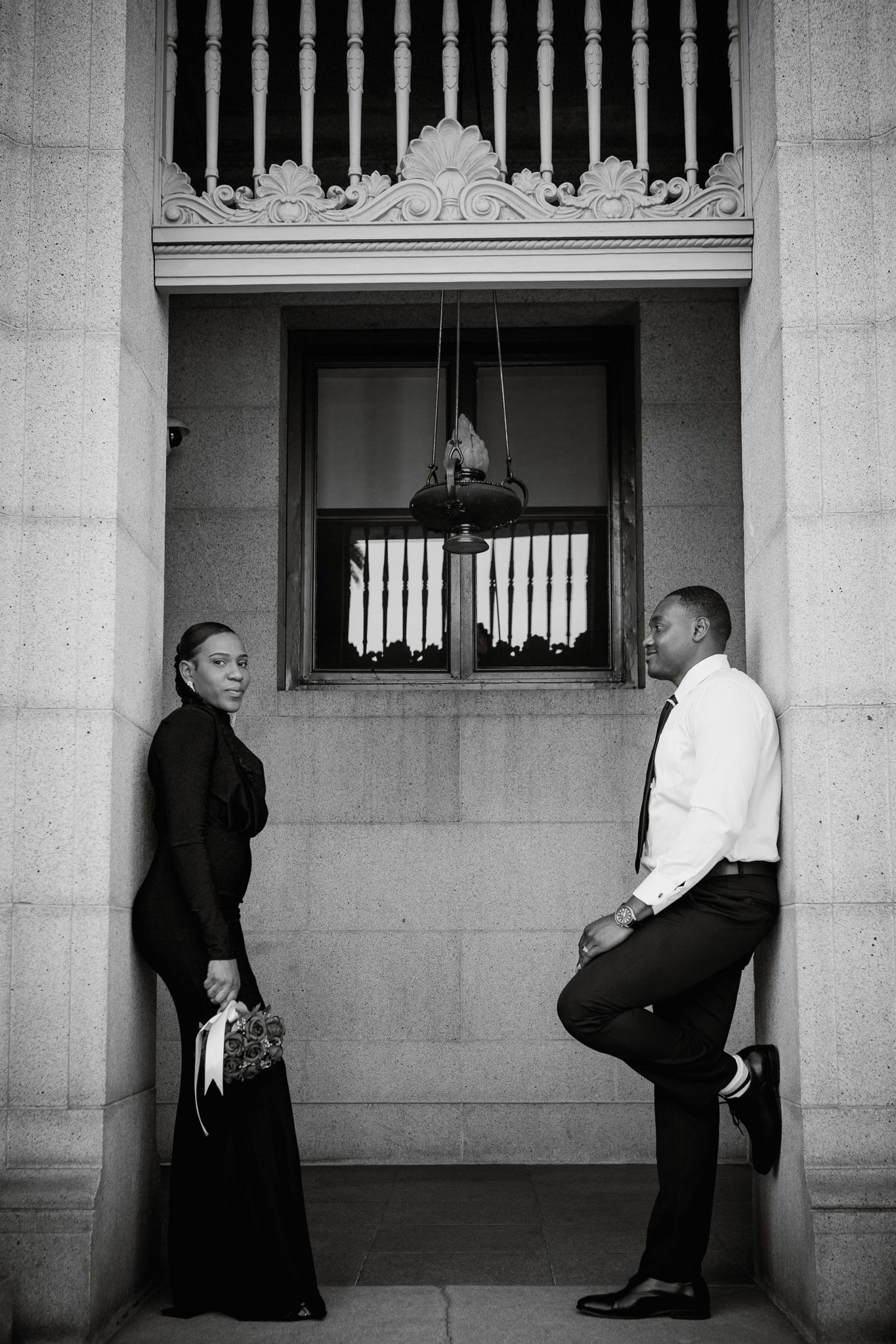 A black and white artistic photograph showing people waiting in an architectural colonnade with dramatic lighting.