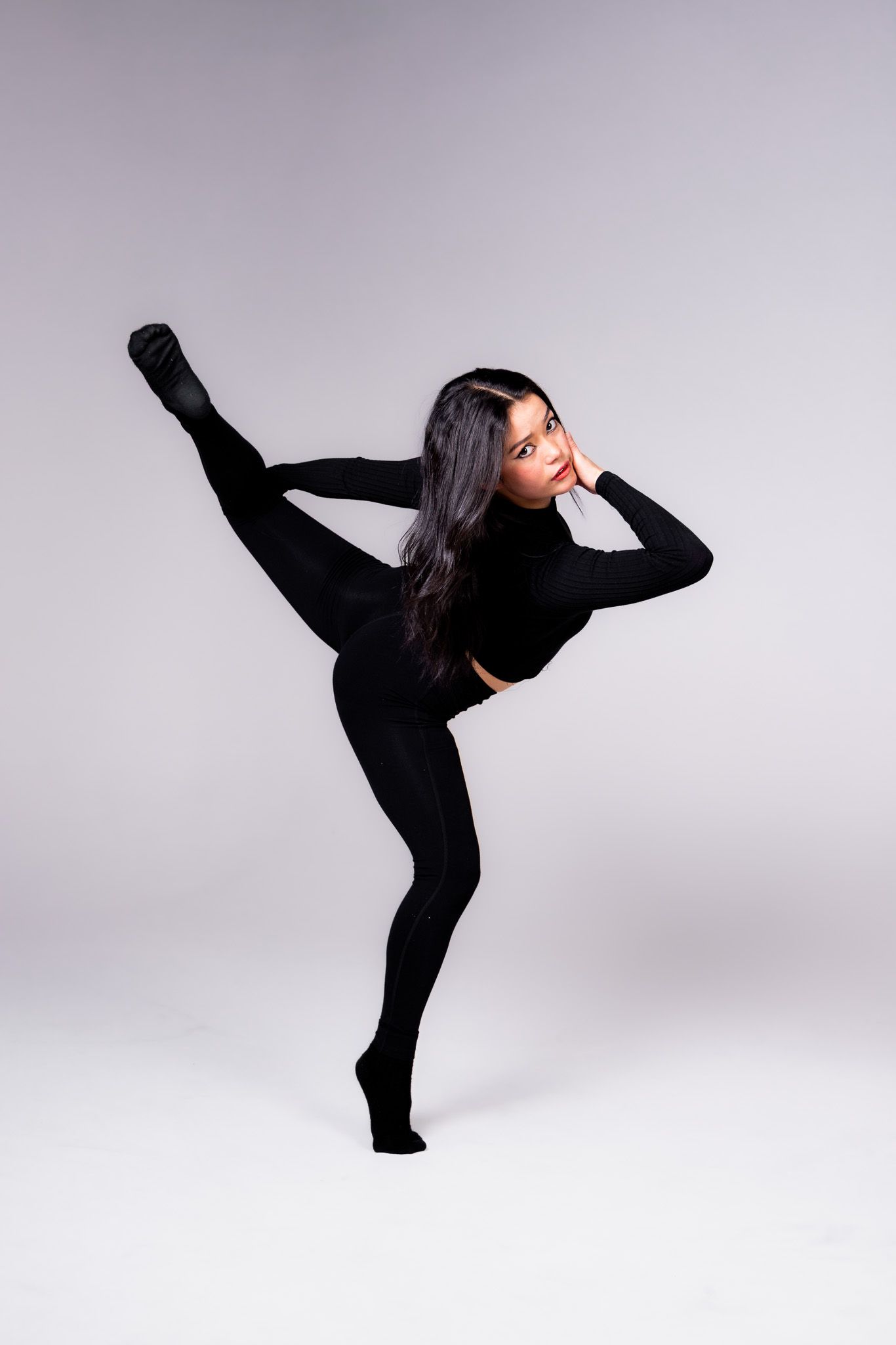 A dancer in black attire performs a dynamic high kick sequence against a light gray studio backdrop.