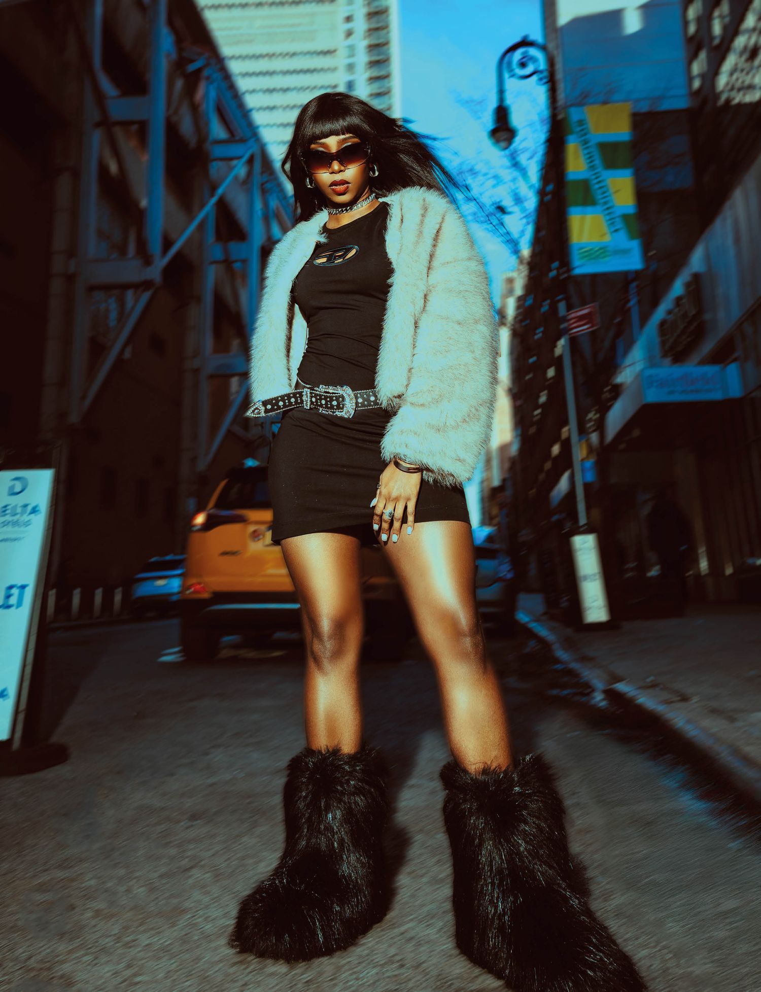 Fashion model wearing furry black boots and dark outfit poses against urban alley backdrop.