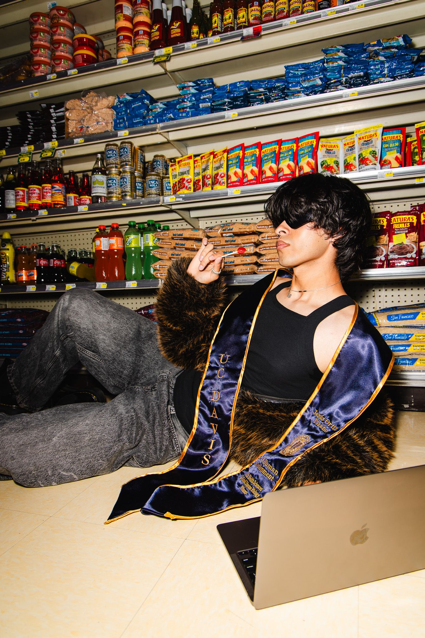 A person in a blue jacket leans back in a chair against a store shelf stocked with snacks and beverages.