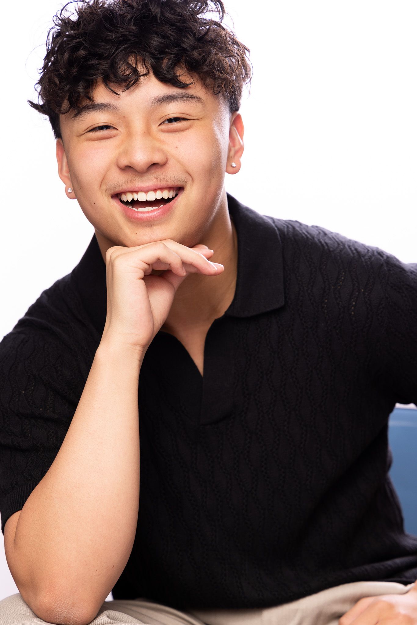 Person in black shirt smiles warmly while resting head on hand against light background.