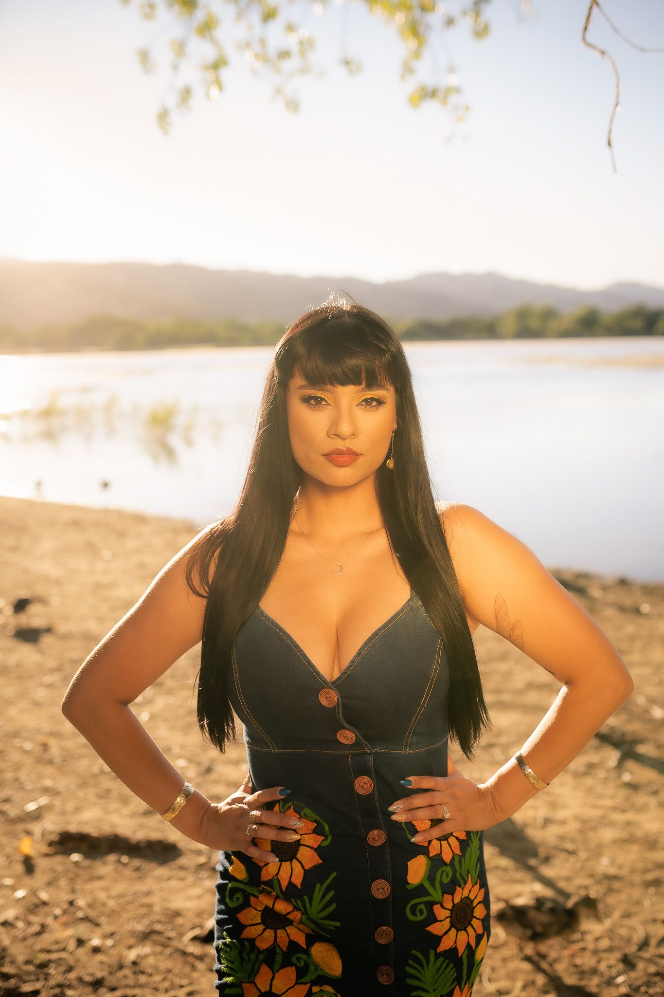 A woman poses outdoors in a black floral dress by the water during golden hour.