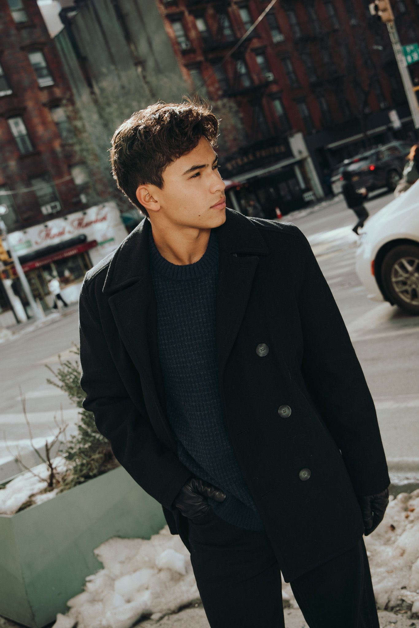 A person in a black coat stands on an urban street with brick buildings in the background.