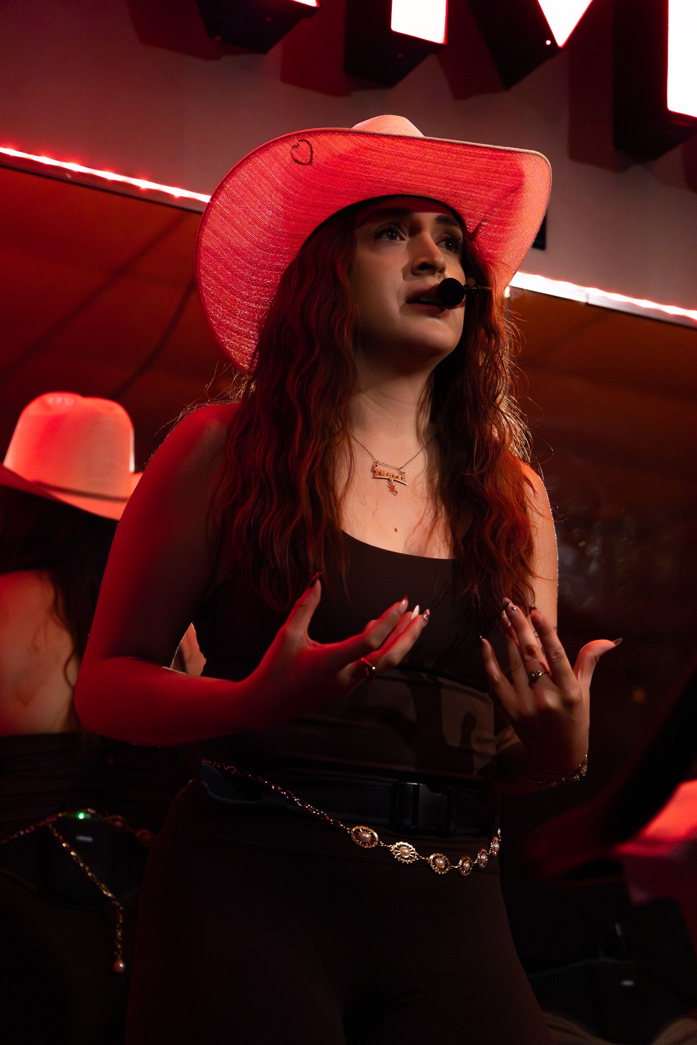 Woman in black dress and red hat poses dramatically under moody red lighting in a dark setting.