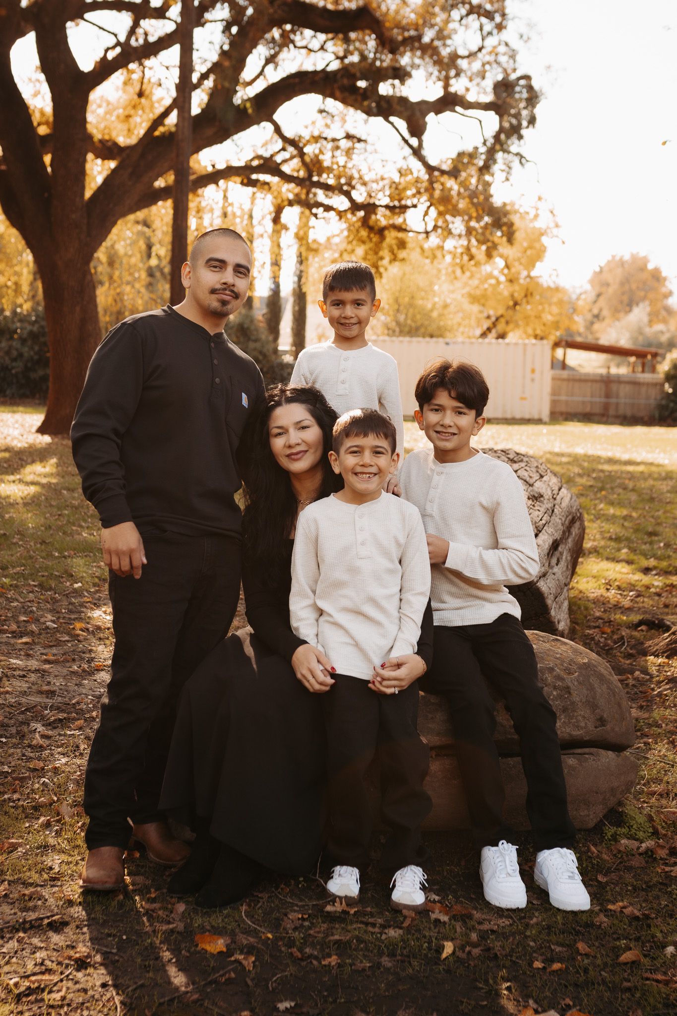 A family sits together on a large rock during a golden hour autumn photoshoot in a park setting.