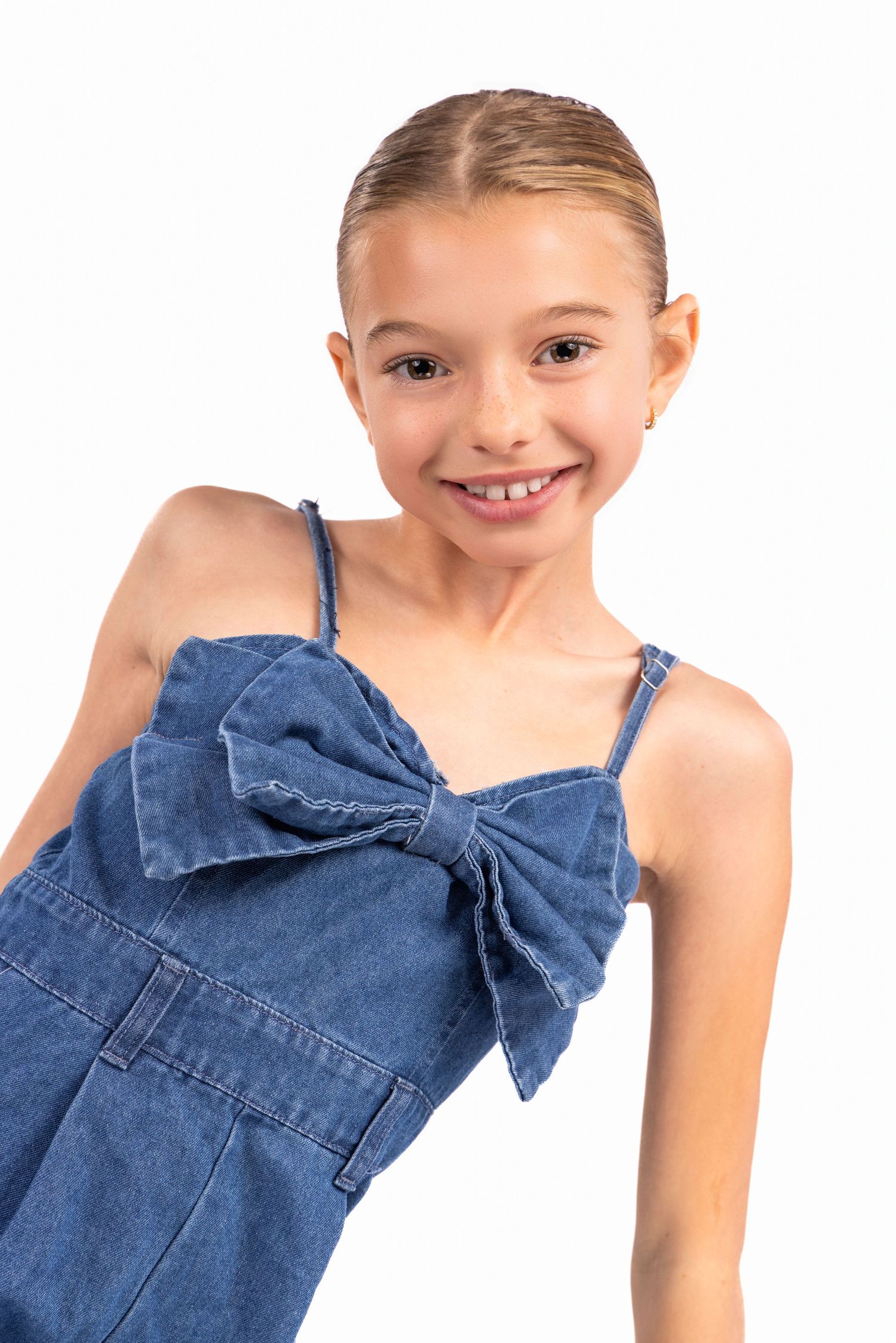 Young person wearing a denim dress with a large bow detail on the bodice against white background.