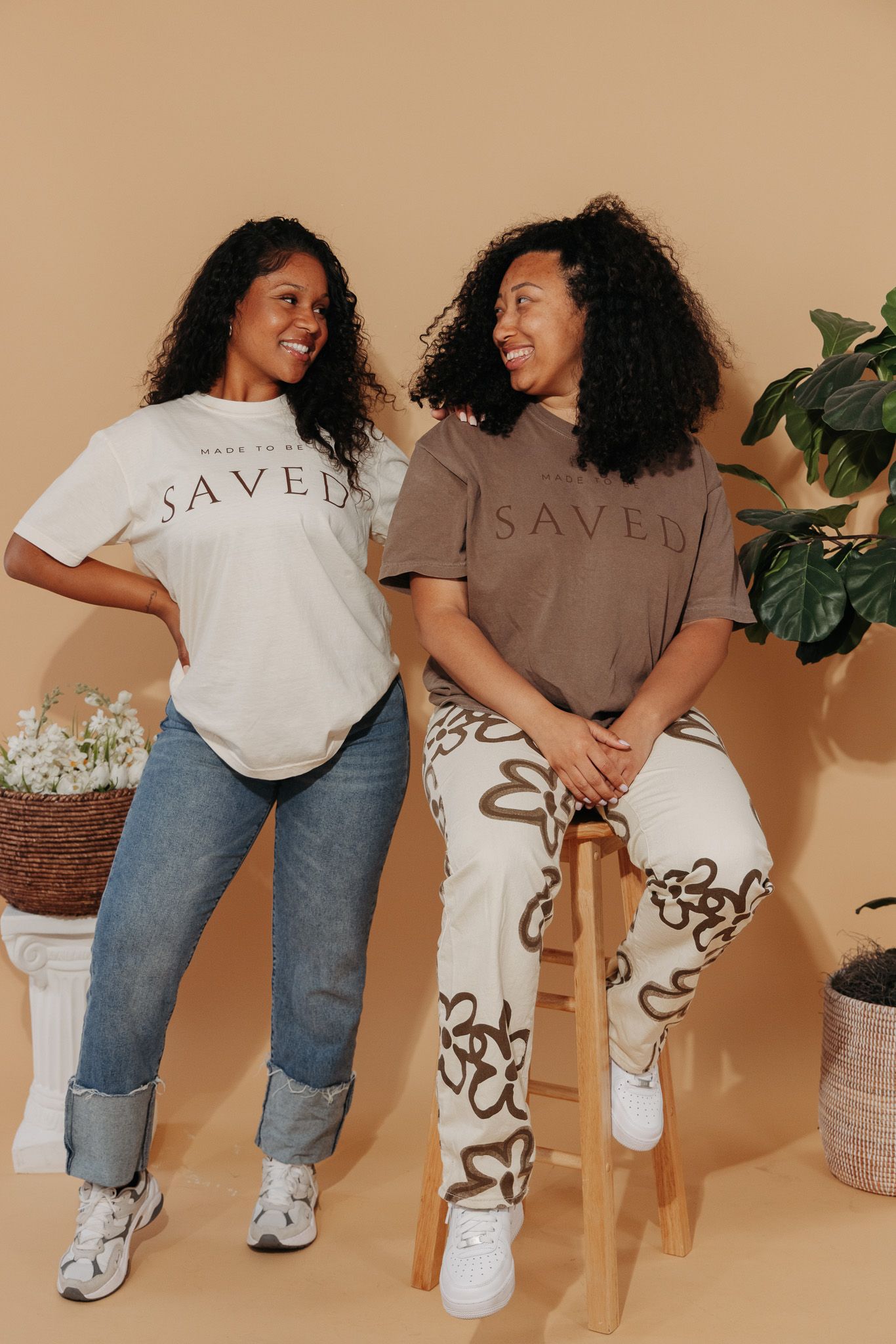 Two friends in casual wear sporting branded t-shirts and jeans pose together with plants in background.