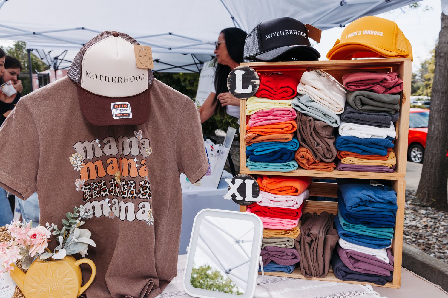 Outdoor market booth displaying Motherhood branded hats, mama graphic tees, and colorful folded shirts on a wooden shelf.