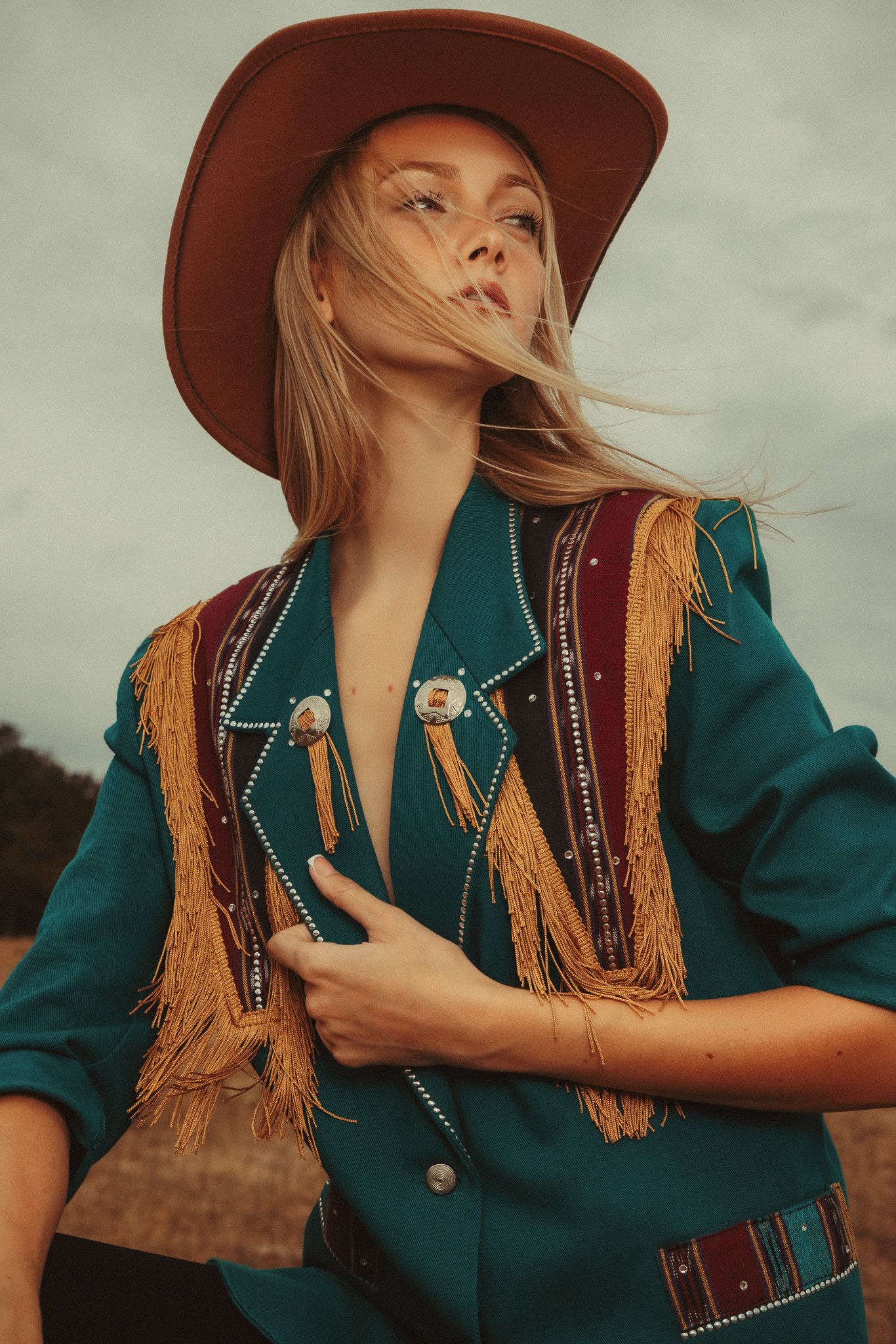 Fashion portrait of a model in a teal western jacket and brown hat posing against a neutral background.