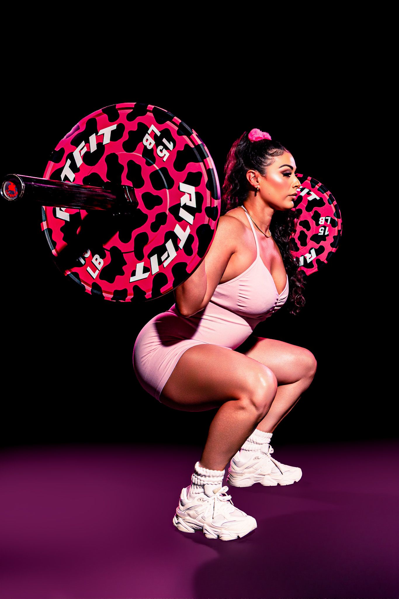 Woman performing a barbell back squat in pink workout outfit with leopard print weights on black background.
