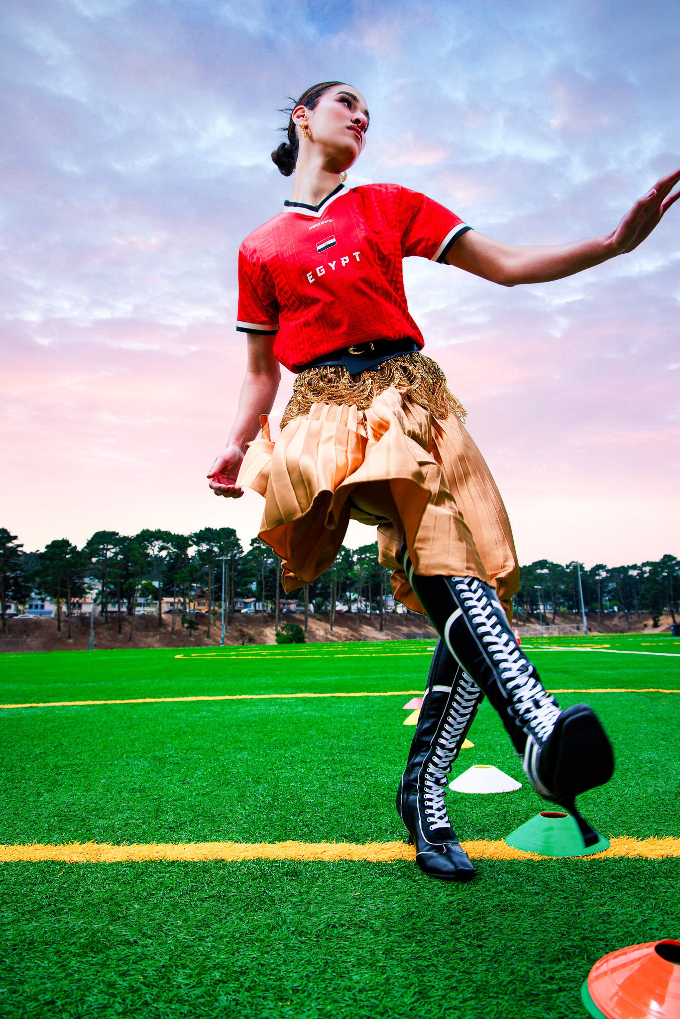 Woman in red Egypt soccer jersey, gold skirt, and black knee-high heels poses dynamically on a green turf field.
