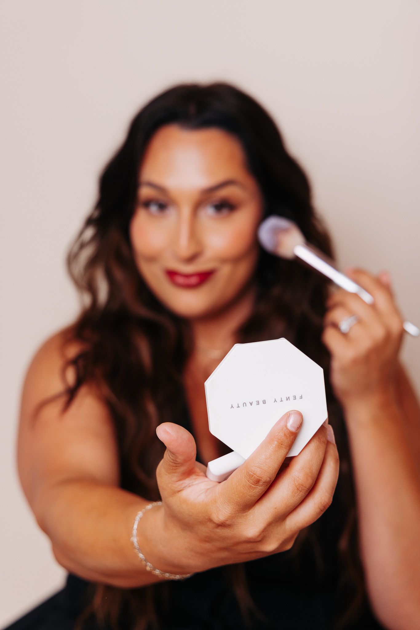 Woman holding Fenty Beauty compact and brush, applying makeup with a soft smile against a neutral background.