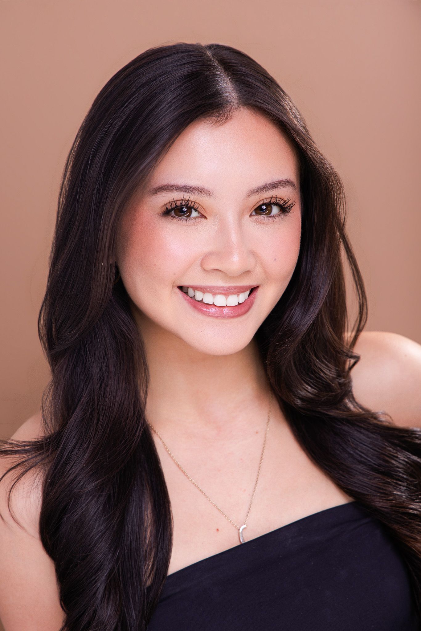 Smiling young woman with long dark hair wearing a black strapless top and delicate necklace against a brown background.