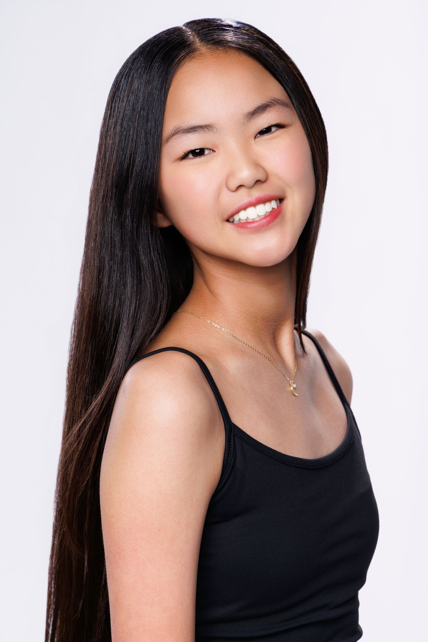Smiling young Asian woman with long black hair wearing a black tank top and gold necklace against white background.