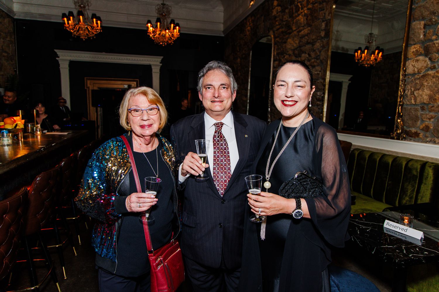 Three elegantly dressed guests smile and hold drinks at an upscale bar event.