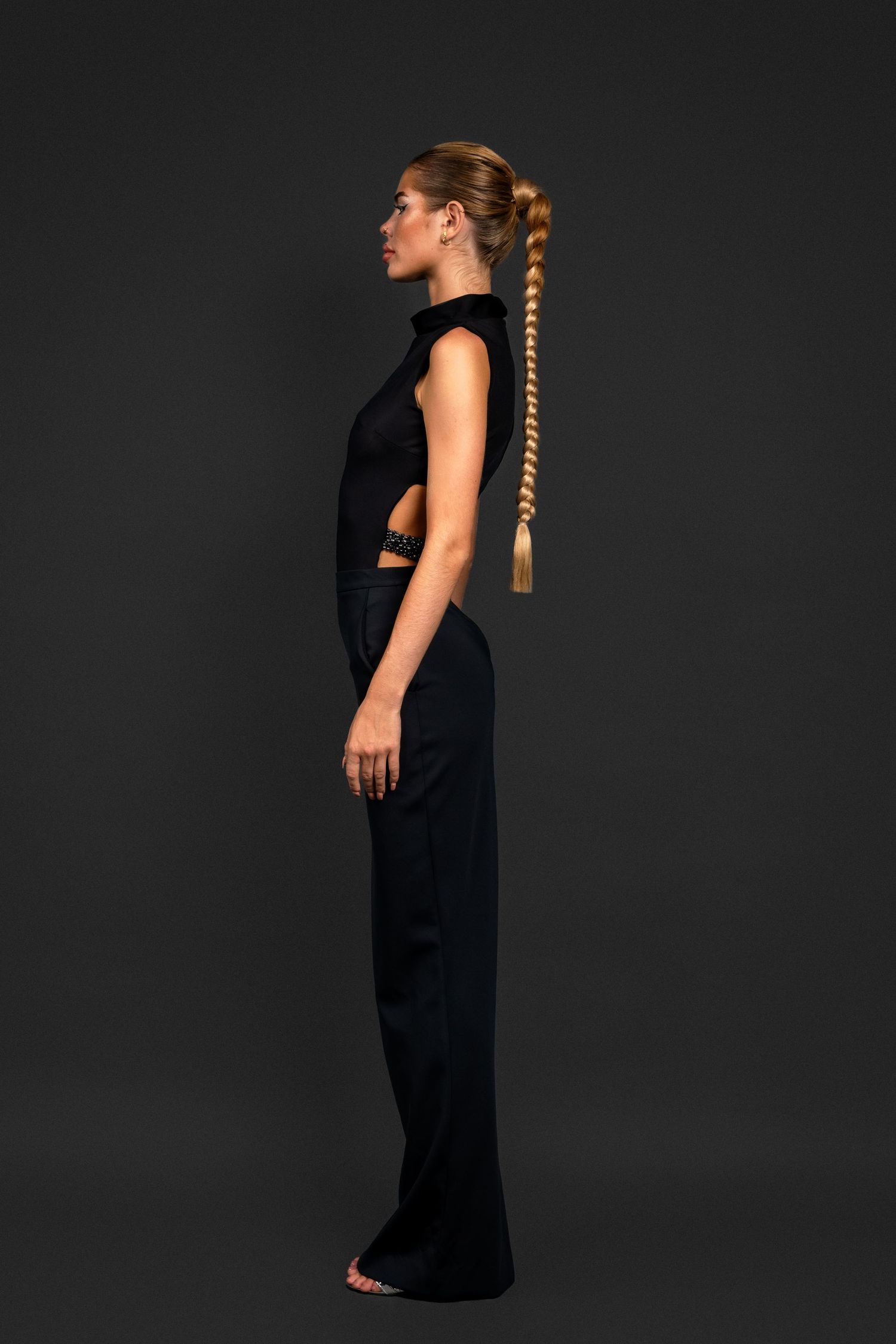 Model in sleek black sleeveless cutout jumpsuit with long braided ponytail, posing in profile against dark background.