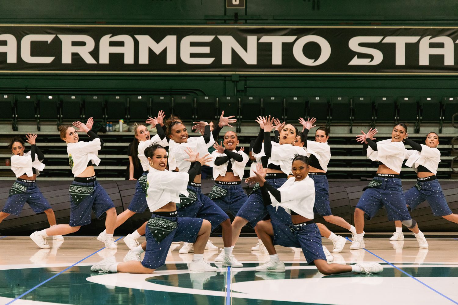 Sacramento State dance team performing synchronized routine in white tops and dark skirts on a green basketball court.