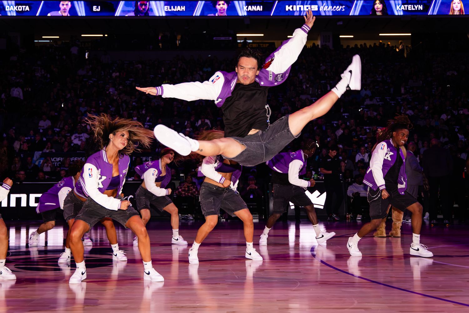 Energetic NBA halftime dancers performing acrobatic moves on a purple-lit arena court during a basketball game.