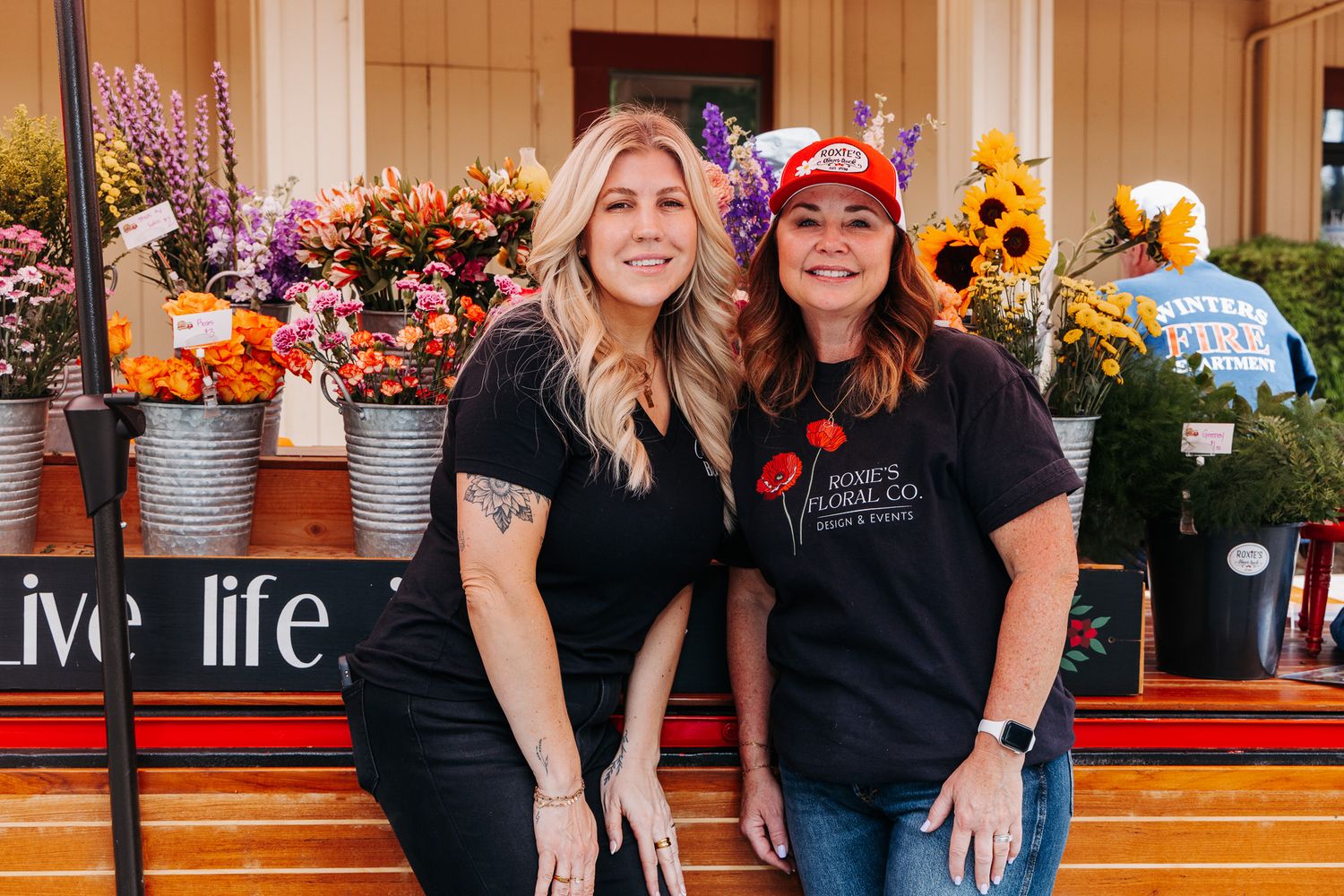 Two women in black t-shirts smile in front of a colorful flower market display with sunflowers and mixed blooms.