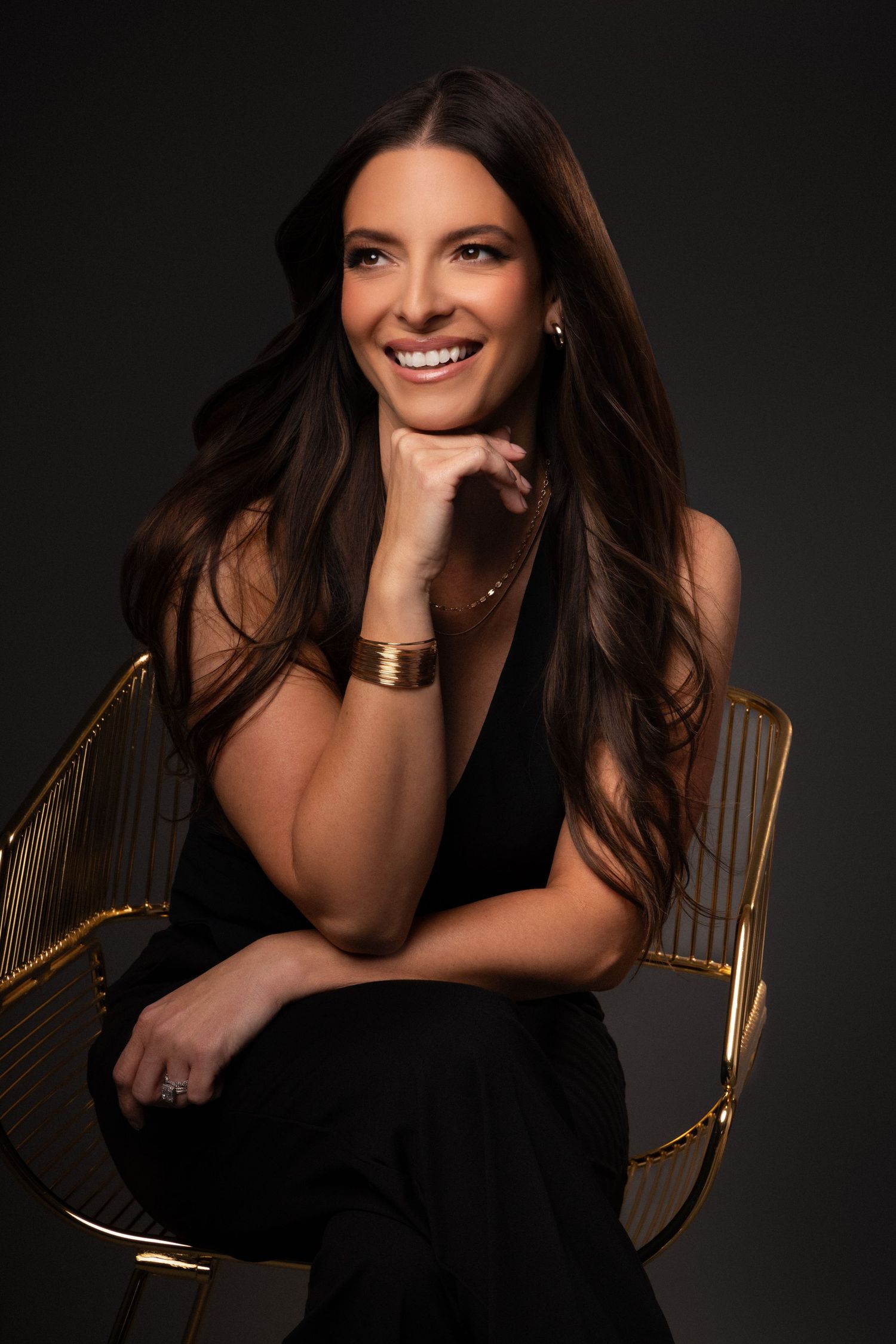 Professional portrait of a woman in a black outfit sitting in a gold chair with a warm smile against dark background.