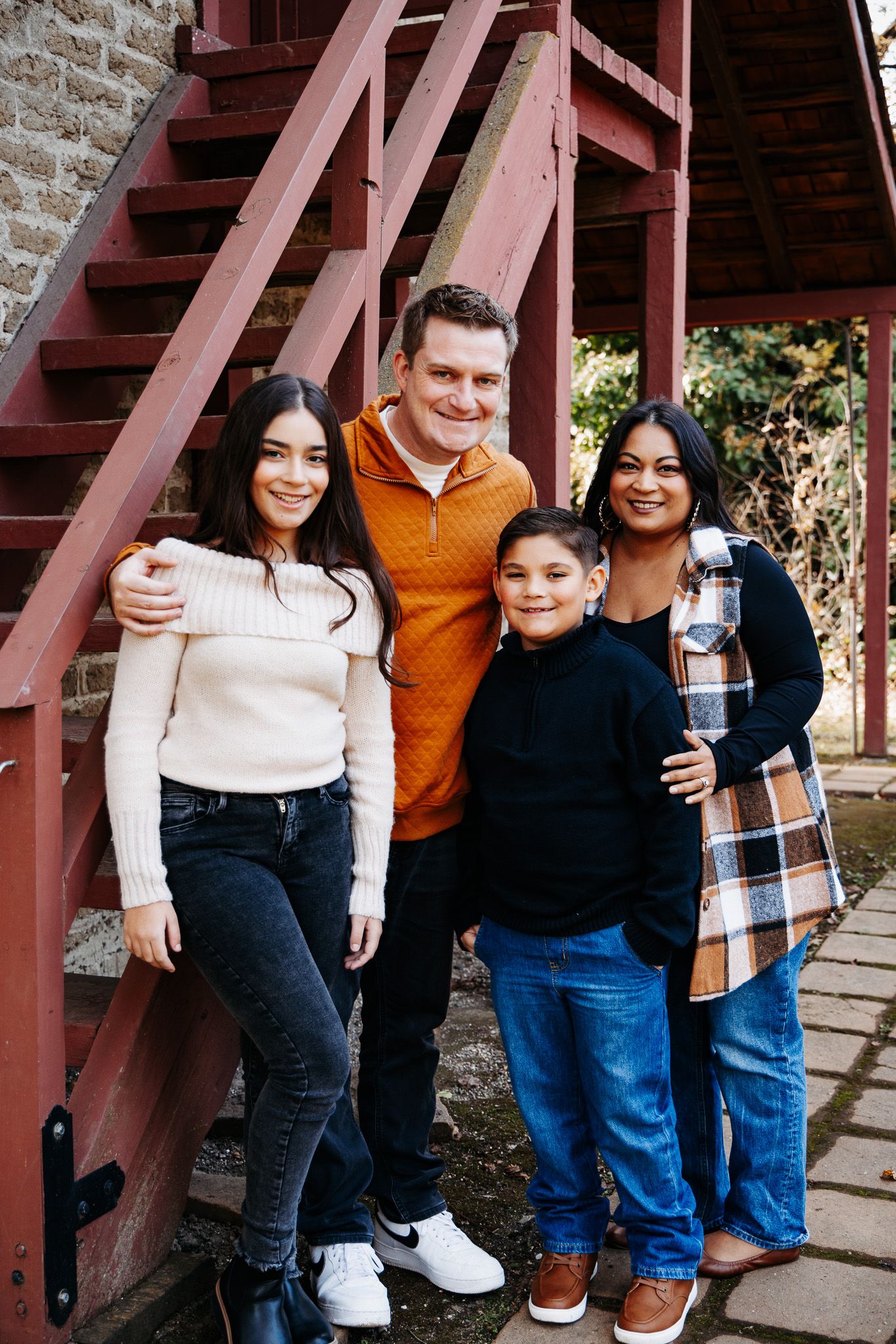 A group stands together smiling on red wooden stairs outside a stone building on a sunny day.